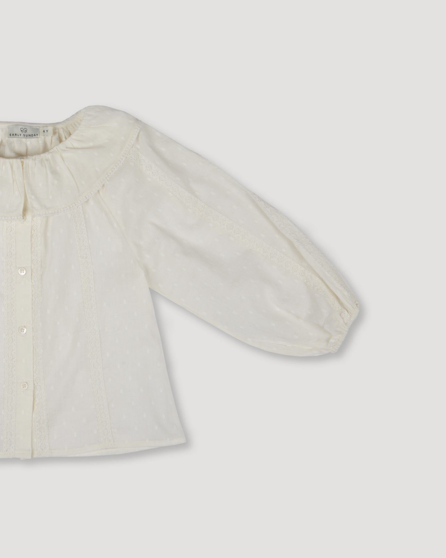 Early Sunday, Cecilia Blouse in Ecru color, made from Cotton Swiss Dobby fabric, featuring relaxed fit, frill neckline, long sleeves with elasticated cuffs, and MOP buttons, shown in a closeup flat-lay view