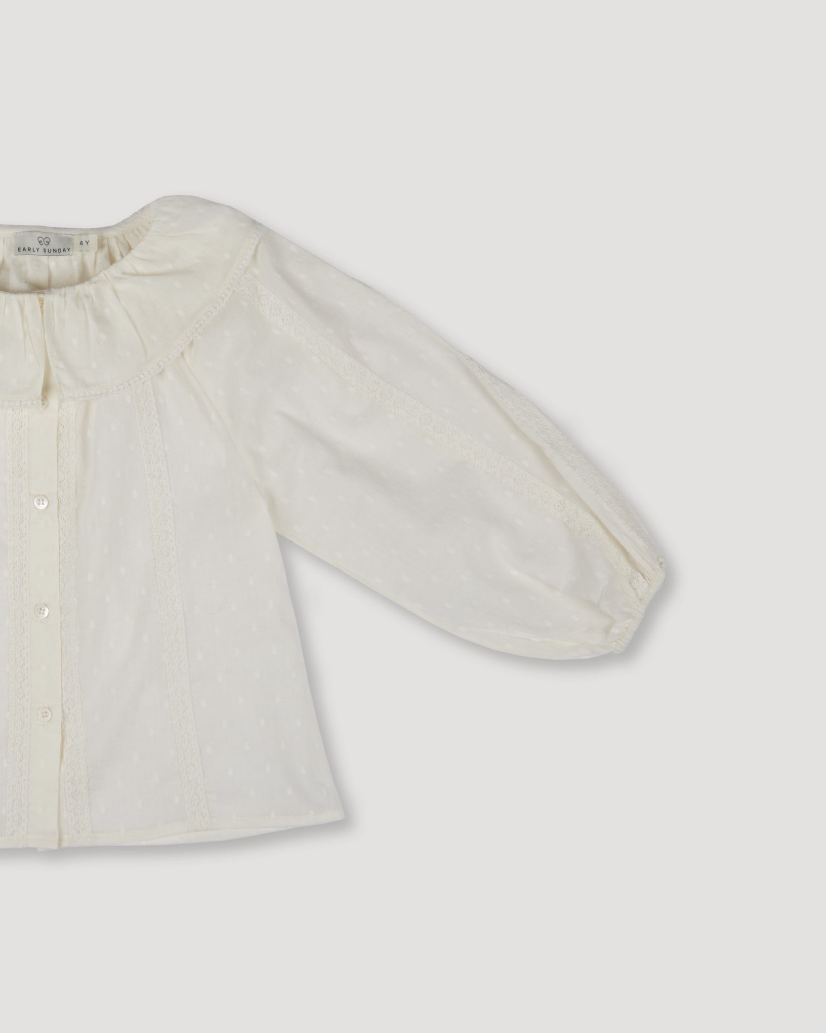 Early Sunday, Cecilia Blouse in Ecru color, made from Cotton Swiss Dobby fabric, featuring relaxed fit, frill neckline, long sleeves with elasticated cuffs, and MOP buttons, shown in a closeup flat-lay view