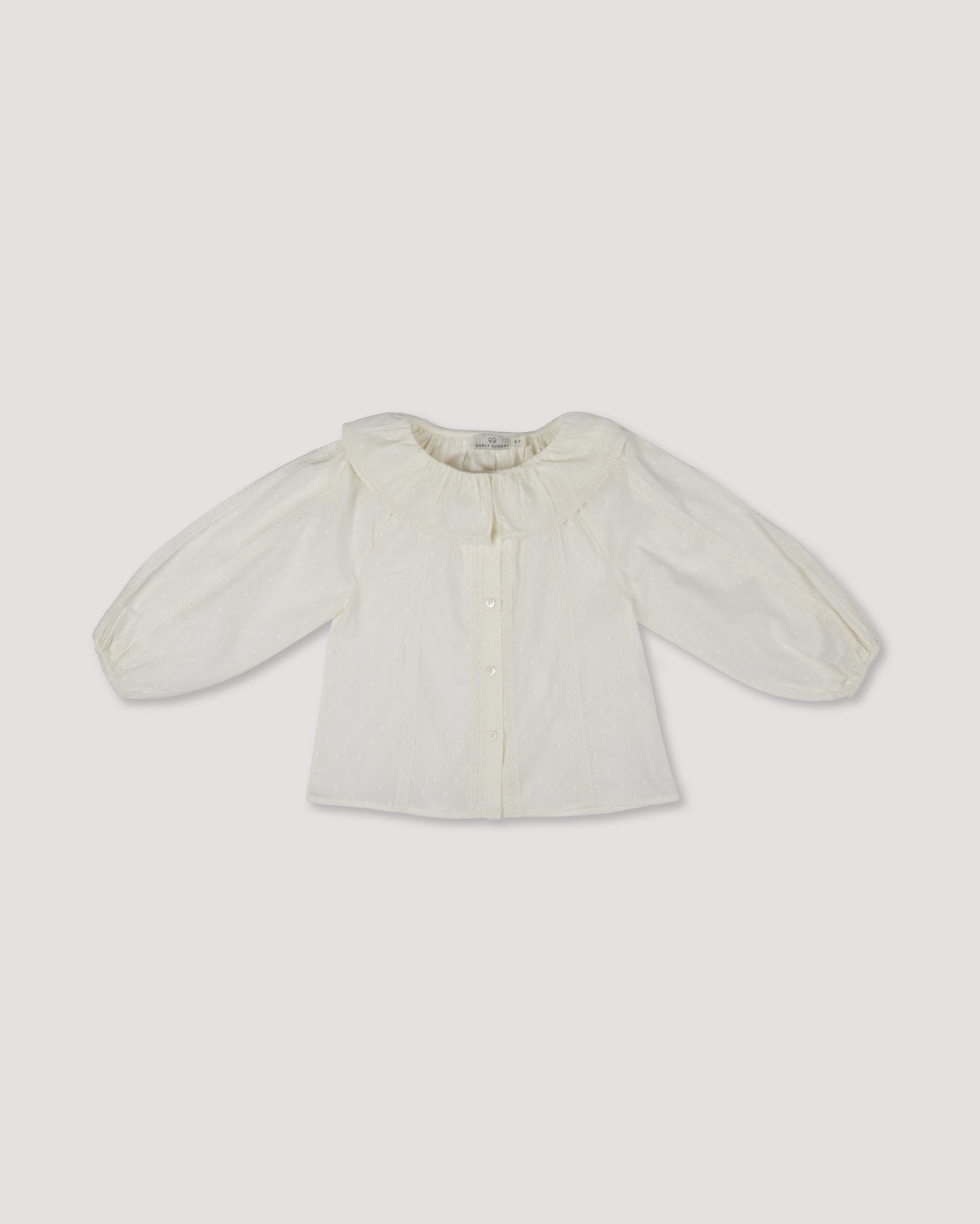 Early Sunday, Cecilia Blouse in Ecru print, made from Cotton Swiss Dobby fabric, featuring relaxed fit, frill neckline, long sleeves with elasticated cuffs, and MOP buttons, shown in a front flat-lay view
