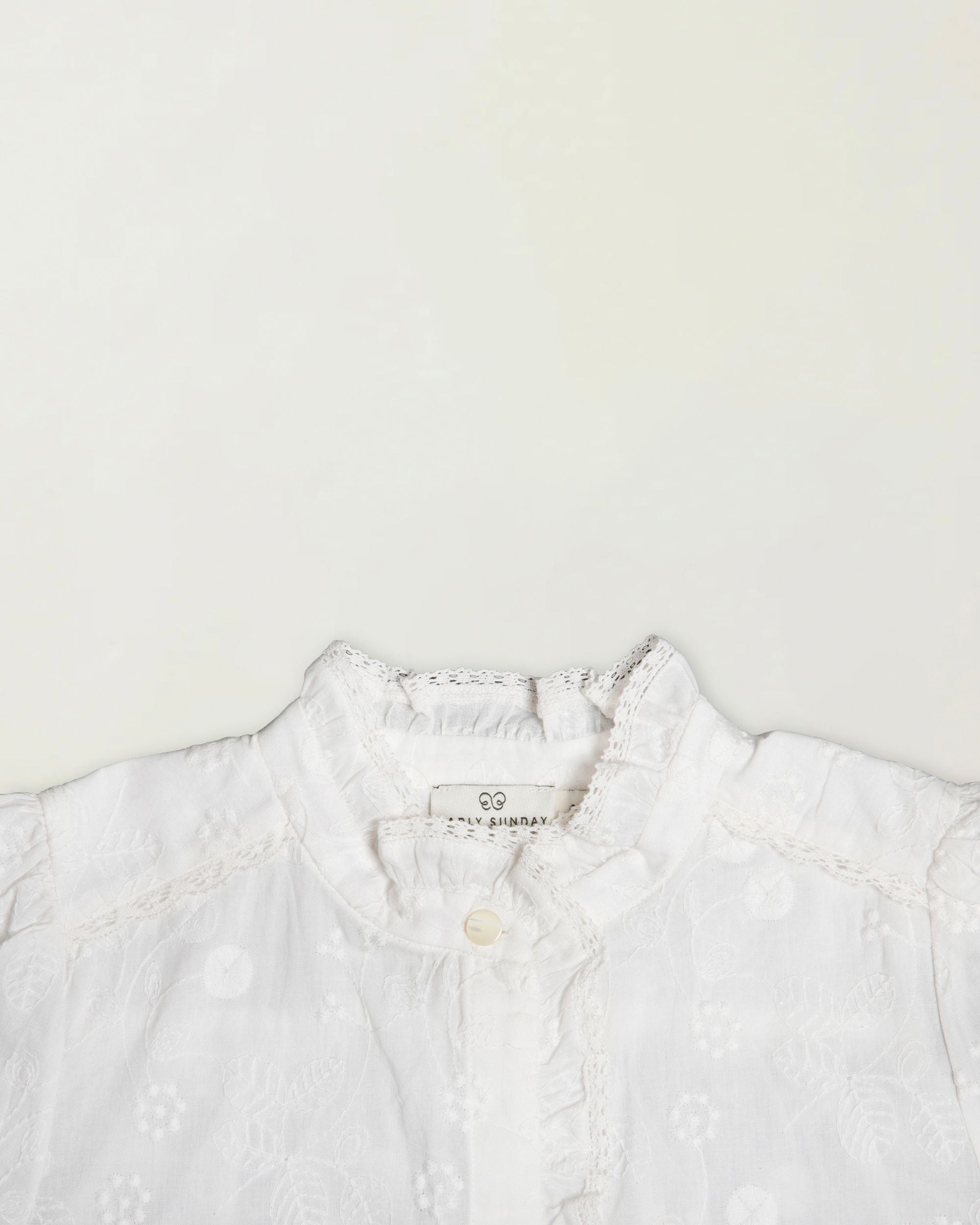 Early Sunday, Coco Blouse in Ivory color, made from all over embroidery fabric, featuring Puff sleeves, ruffled neckline and placket, shown in a closeup flat-lay view