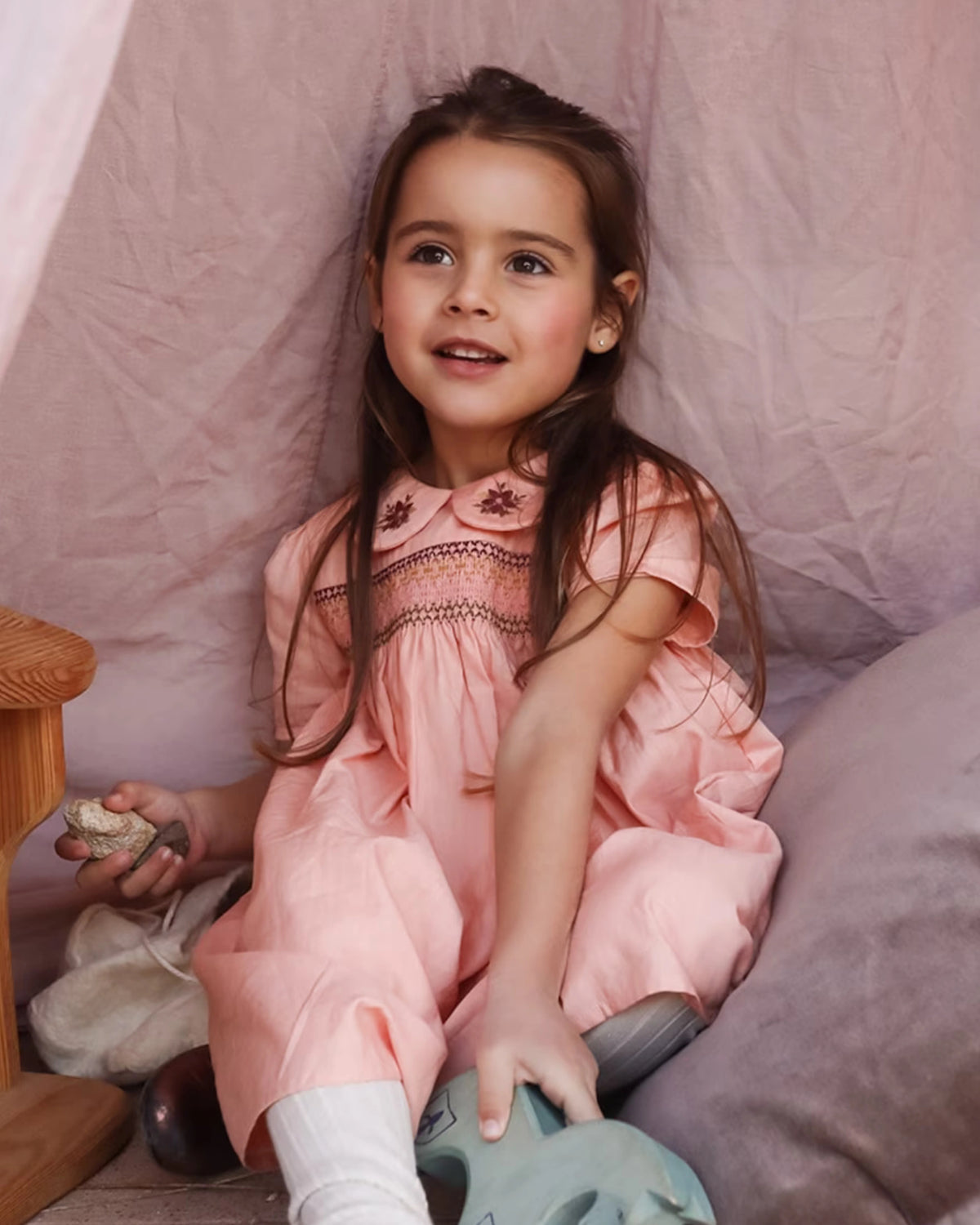 Early Sunday, Erika Dress in Blush Pink, made from Cotton Satin fabric, featuring a floral embroidery on a Peter Pan collar with a delicately smocked chest, shown in a front view of a seated model during a photo shoot