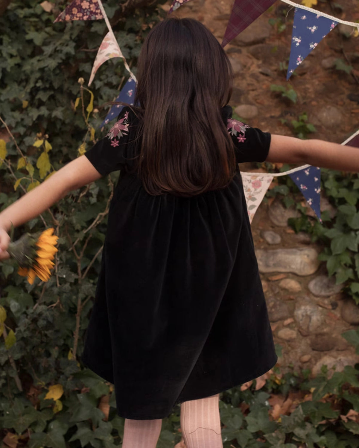 Early Sunday, Erika Dress in Black, made from Cotton Velvet Velour's fabric, featuring a beautiful embroidery with frilled neckline, shown in a back view of model wearing outdoors