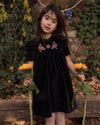 Early Sunday, Erika Dress in Black, made from Cotton Velvet Velour's fabric, featuring a beautiful embroidery with frilled neckline, shown in a front view of model wearing outdoors