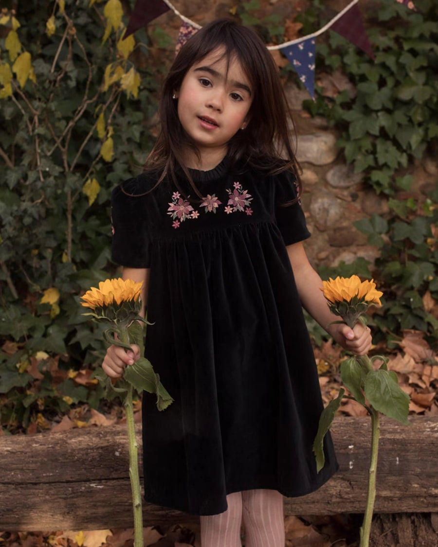 Early Sunday, Erika Dress in Black, made from Cotton Velvet Velour's fabric, featuring a beautiful embroidery with frilled neckline, shown in a front view of model wearing outdoors