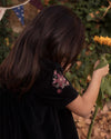 Early Sunday, Erika Dress in Black, made from Cotton Velvet Velour's fabric, featuring a beautiful embroidery with frilled neckline, shown in a side view of model playing outdoors