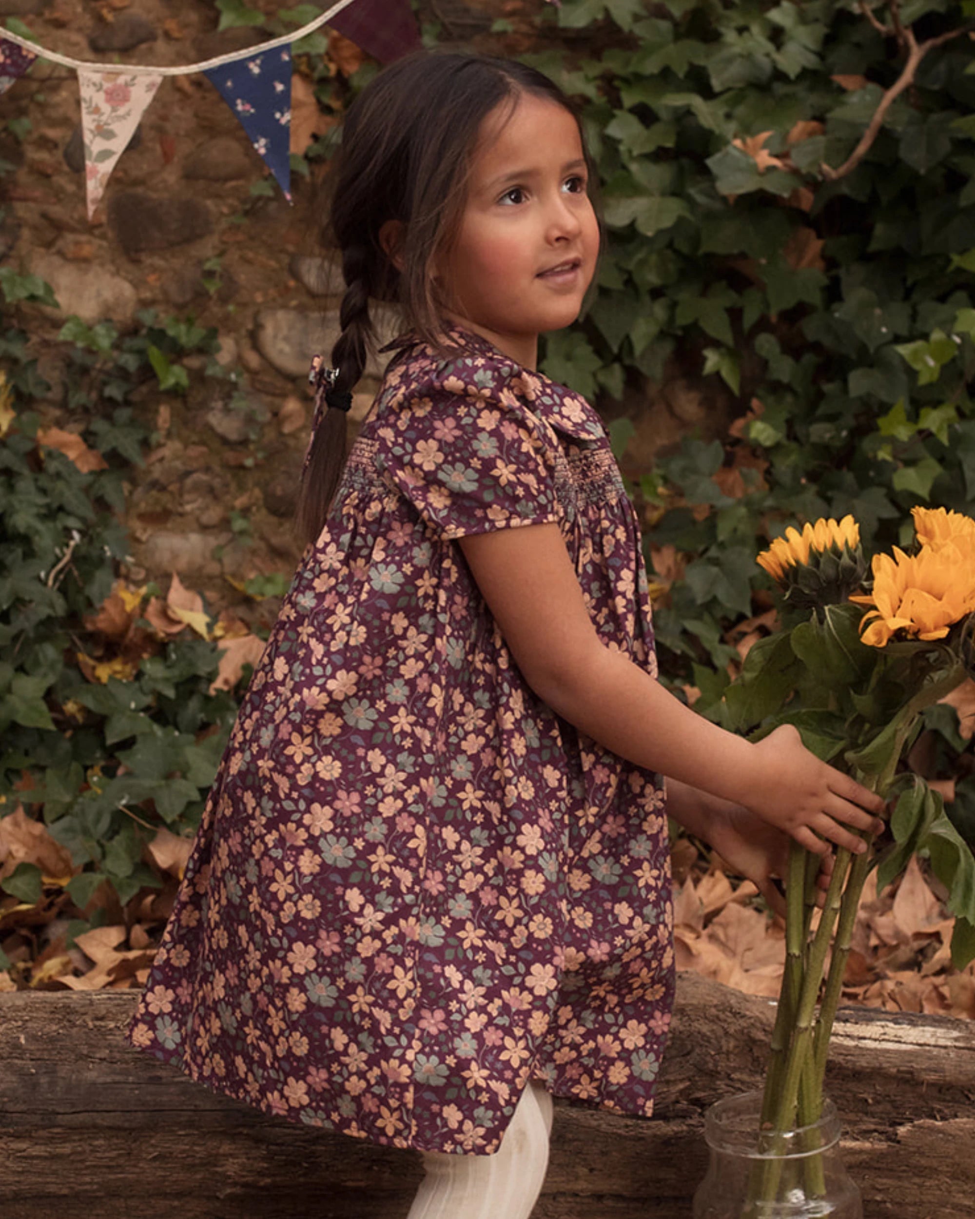 Early Sunday, Erika Dress in Plum, made from Printed Cotton Satin fabric, featuring a floral print with peter pan collar, shown in a side view of model playing outdoors