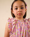 Early Sunday, Florence Dress in Emma Floral Purple color, made from printed cotton cambric fabric, featuring Ruffle detailing on the yoke and sleeves, lace accents, gathered waist with tassel tie, shown in a close up shot of model wearing it