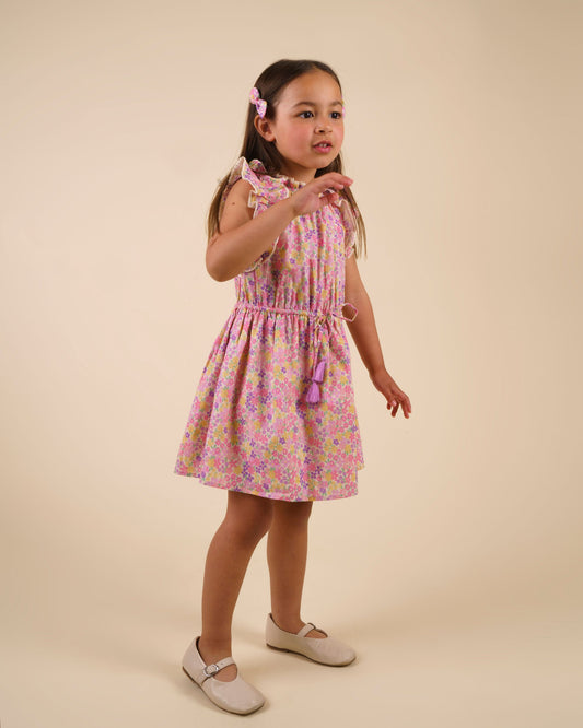 Early Sunday, Florence Dress in Emma Floral Purple color, made from printed cotton cambric fabric, featuring Ruffle detailing on the yoke and sleeves, lace accents, gathered waist with tassel tie, shown in a front view of model wearing