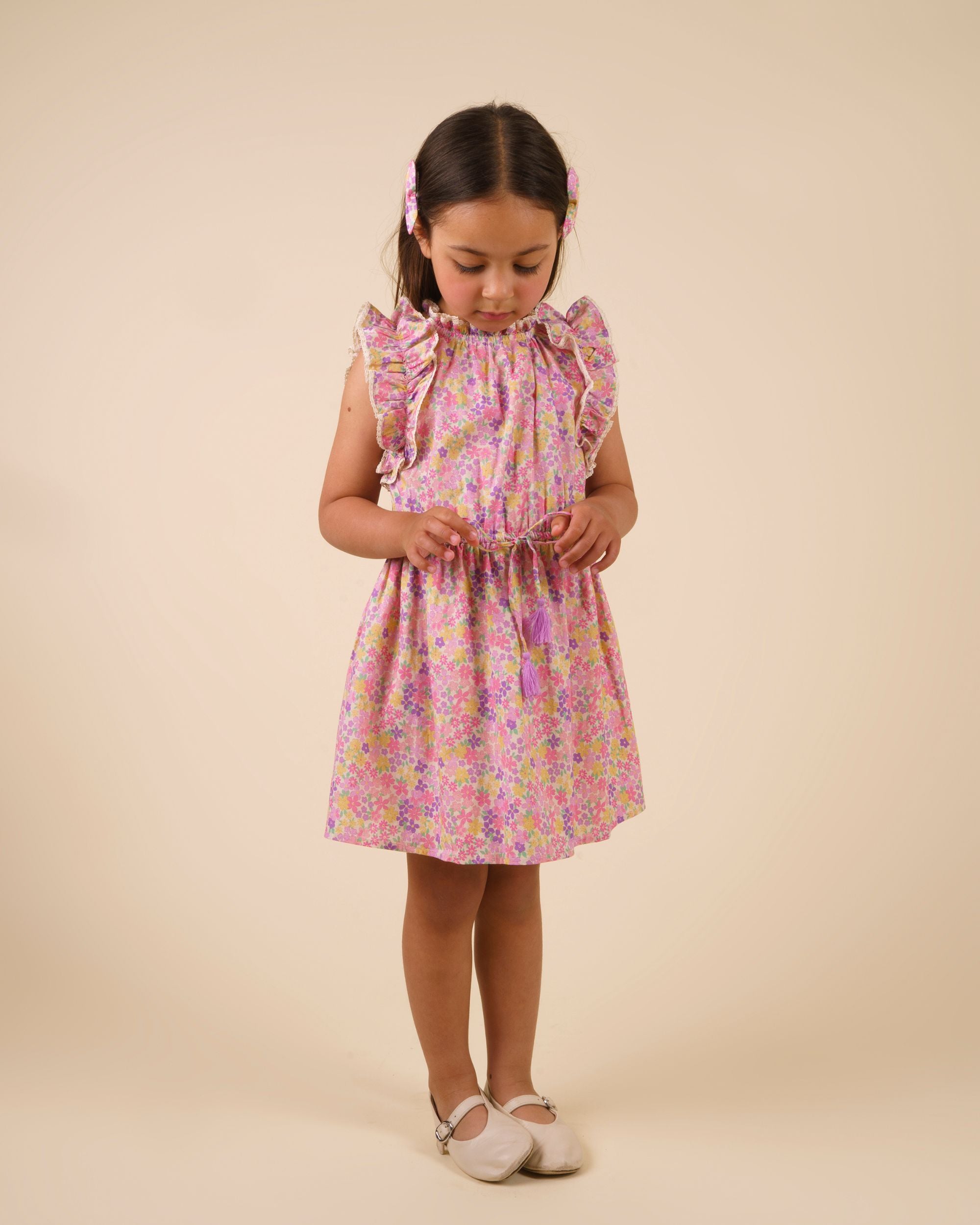 Early Sunday, Florence Dress in Emma Floral Purple color, made from printed cotton cambric fabric, featuring Ruffle detailing on the yoke and sleeves, lace accents, gathered waist with tassel tie, shown in a model standing facing front wearing