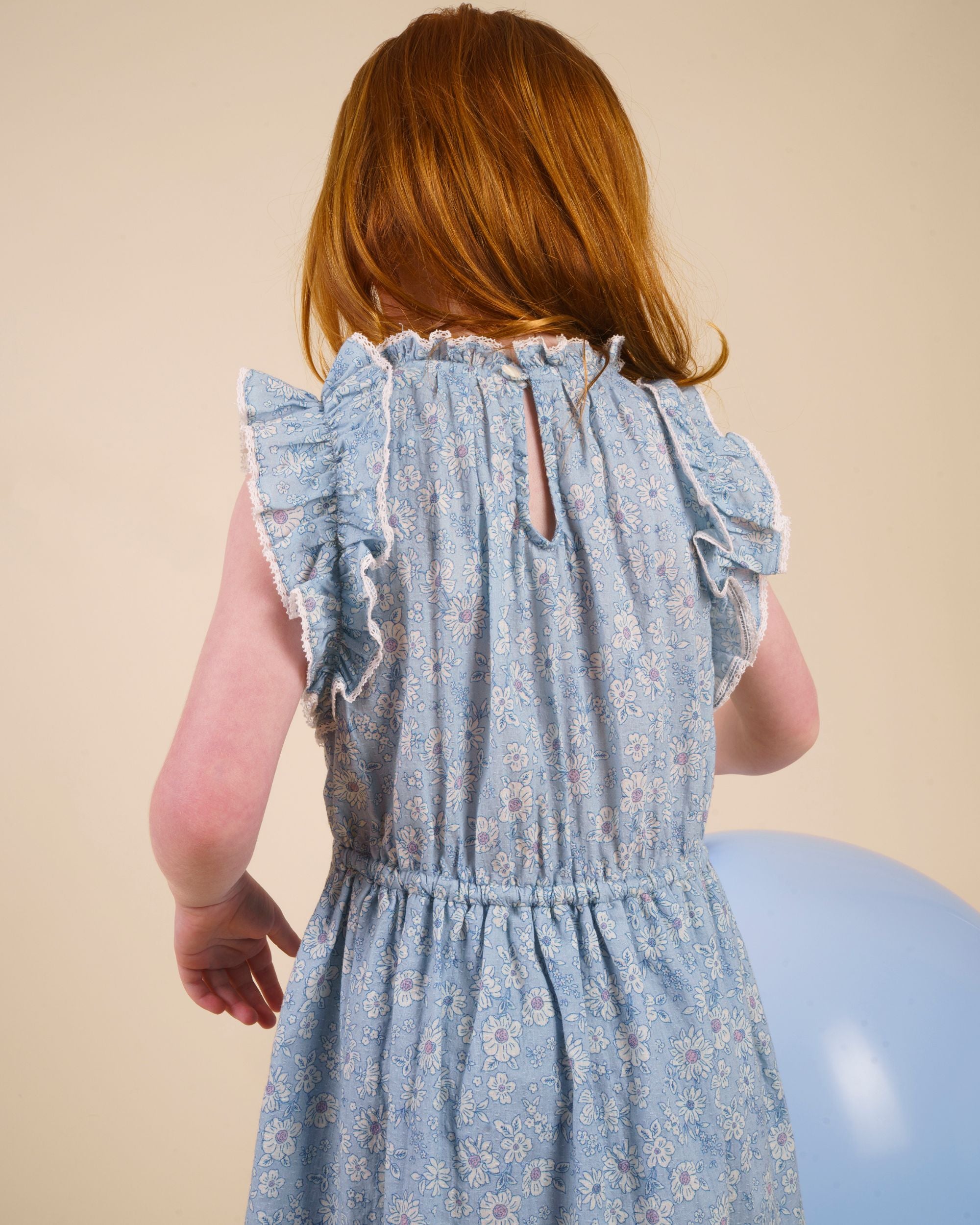 Early Sunday, Florence Dress in Tonal Floral Blue color, made from printed cotton tiny swiss dobby fabric, featuring Ruffle detailing on the yoke and sleeves, lace accents, gathered waist with tassel tie, shown in a back view of model wearing