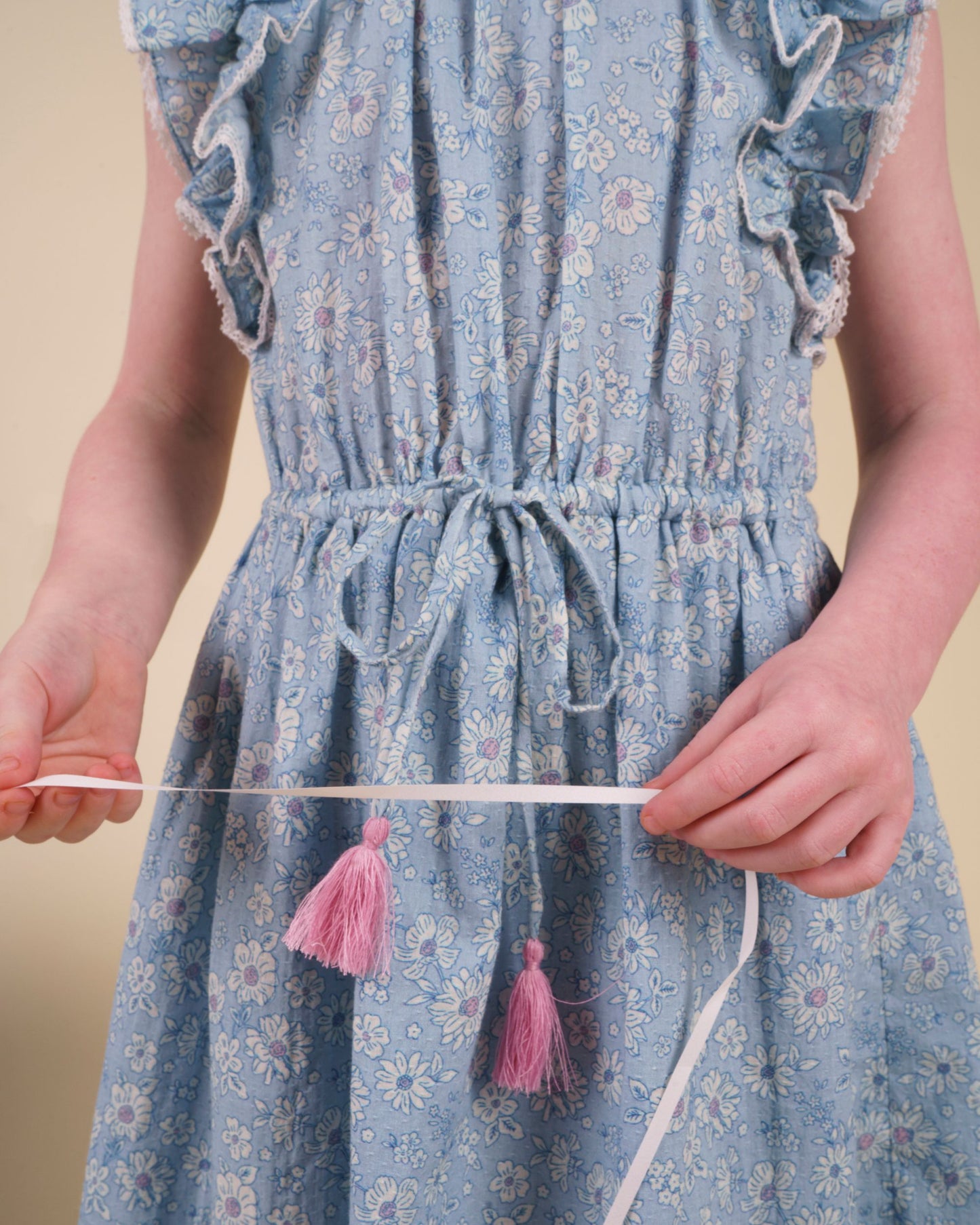 Early Sunday, Florence Dress in Tonal Floral Blue color, made from printed cotton tiny swiss dobby fabric, featuring Ruffle detailing on the yoke and sleeves, lace accents, gathered waist with tassel tie, shown in a close up shot of model wearing it