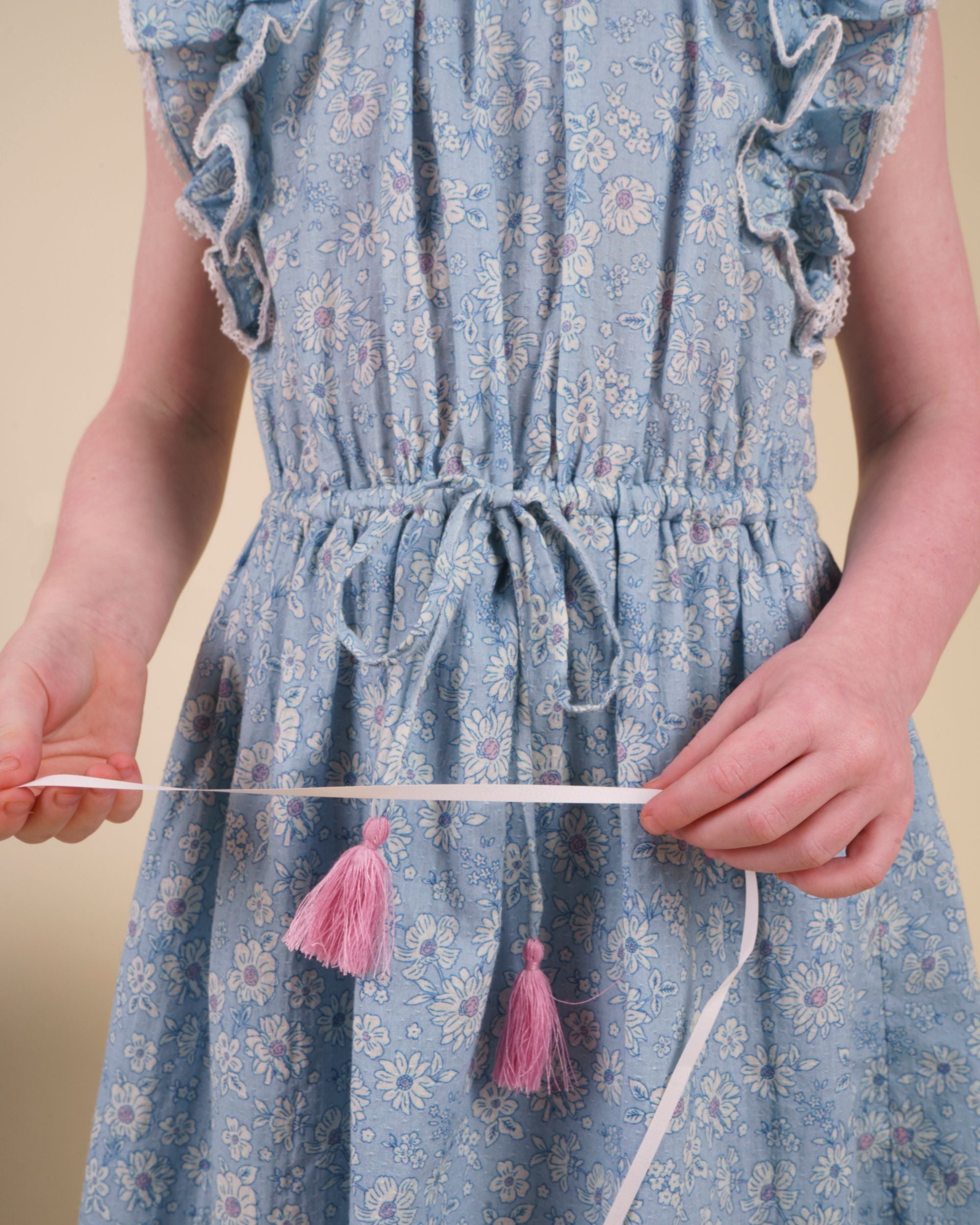 Early Sunday, Florence Dress in Tonal Floral Blue color, made from printed cotton tiny swiss dobby fabric, featuring Ruffle detailing on the yoke and sleeves, lace accents, gathered waist with tassel tie, shown in a close up shot of model wearing it