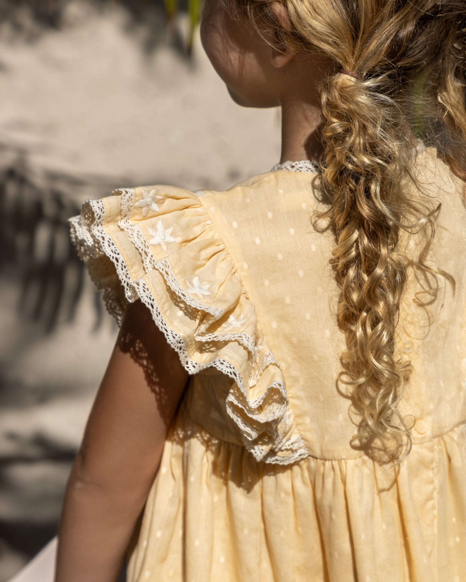 Early Sunday, Georgia Dress in Vanilla color, made from Cotton Dobby Swiss Dots fabric, featuring Flutter sleeves, pintuck detailing, and a flowy, gathered silhouette, shown in a back view of model wearing it