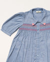 Early Sunday, Jane Dress in Summer Sky color, made from 100% cotton chambray fabric, featuring Smocked / embroidered yoke, peter pan collar and puffed short sleeves, shown in a closeup flat-lay view