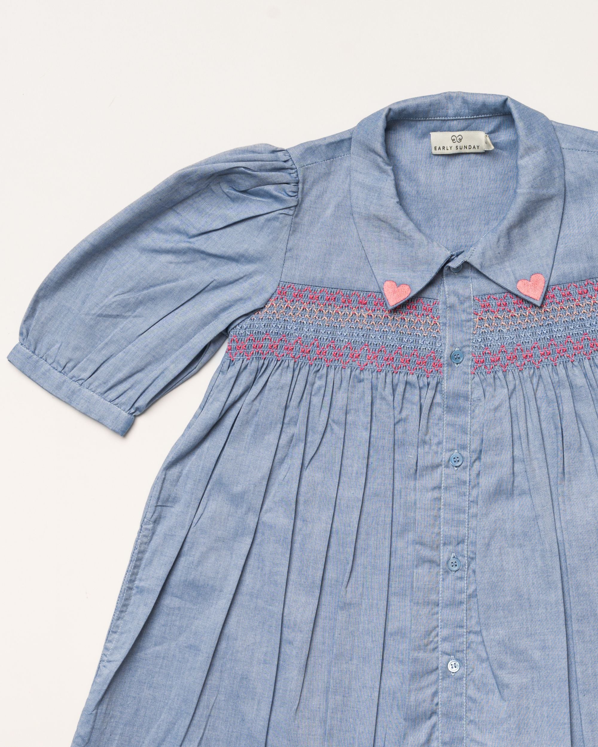 Early Sunday, Jane Dress in Summer Sky color, made from 100% cotton chambray fabric, featuring Smocked / embroidered yoke, peter pan collar and puffed short sleeves, shown in a closeup flat-lay view