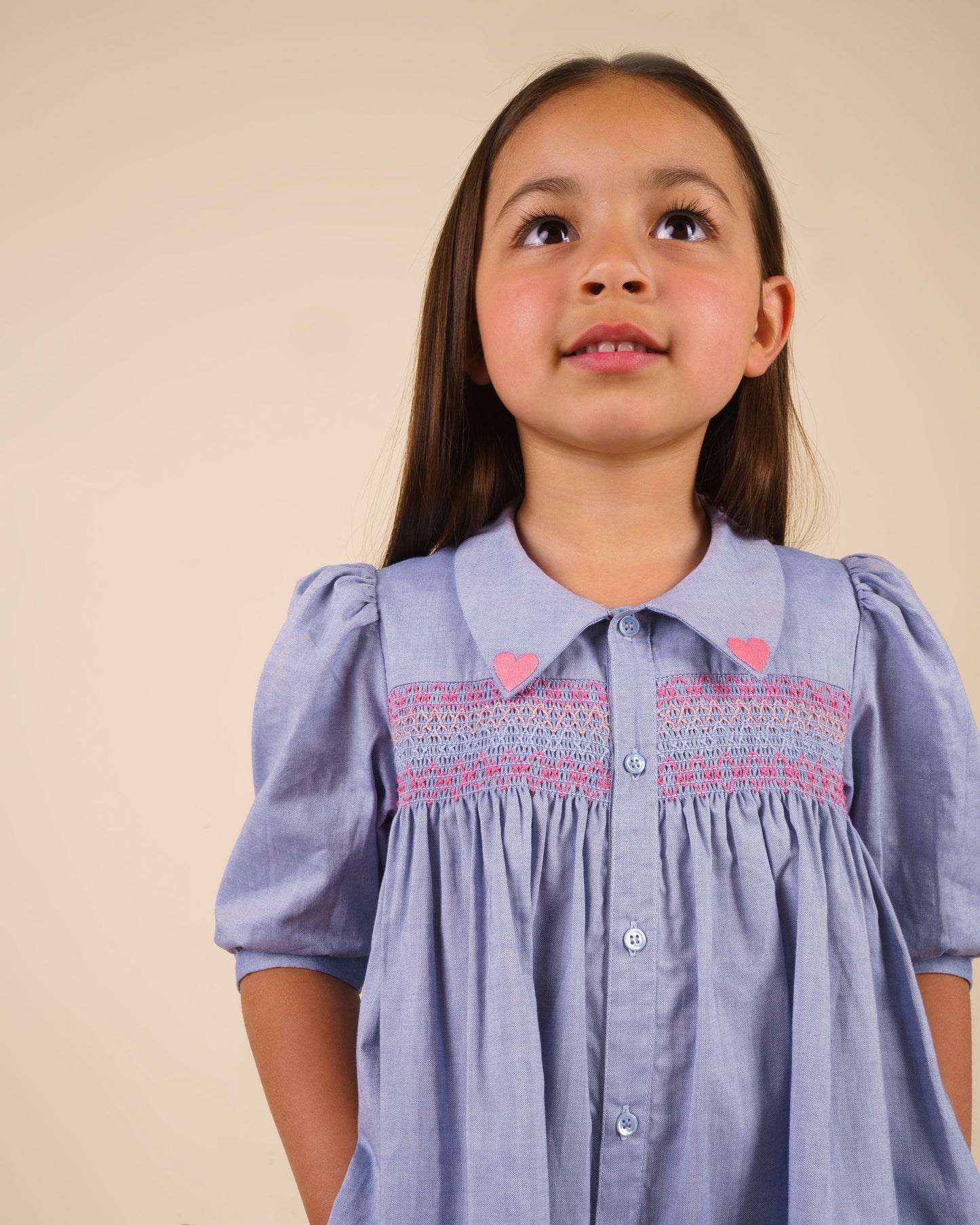 Early Sunday, Jane Dress in Summer Sky color, made from 100% cotton chambray fabric, featuring Smocked / embroidered yoke, peter pan collar and puffed short sleeves, shown in a close up shot of model wearing it