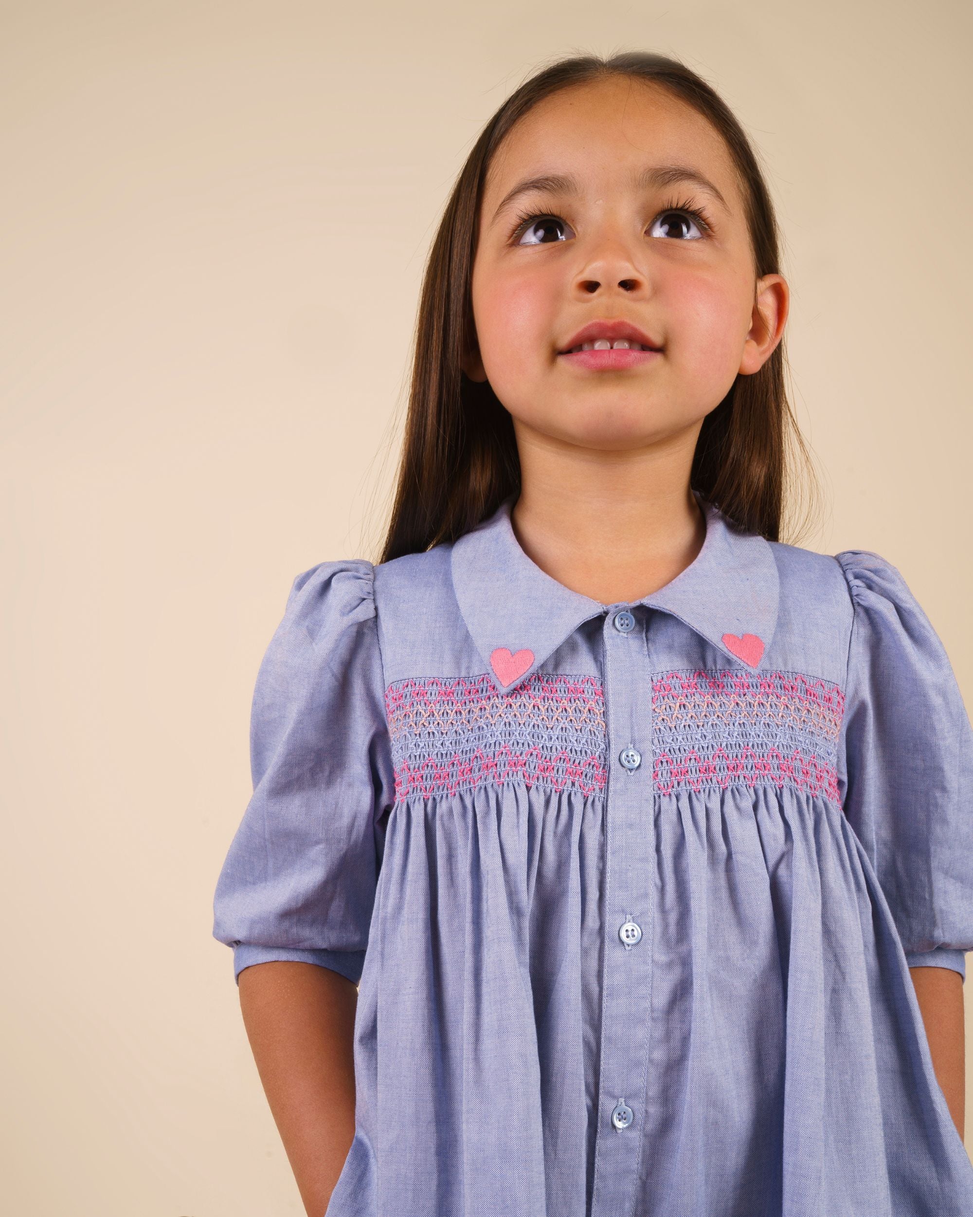 Early Sunday, Jane Dress in Summer Sky color, made from 100% cotton chambray fabric, featuring Smocked / embroidered yoke, peter pan collar and puffed short sleeves, shown in a close up shot of model wearing it