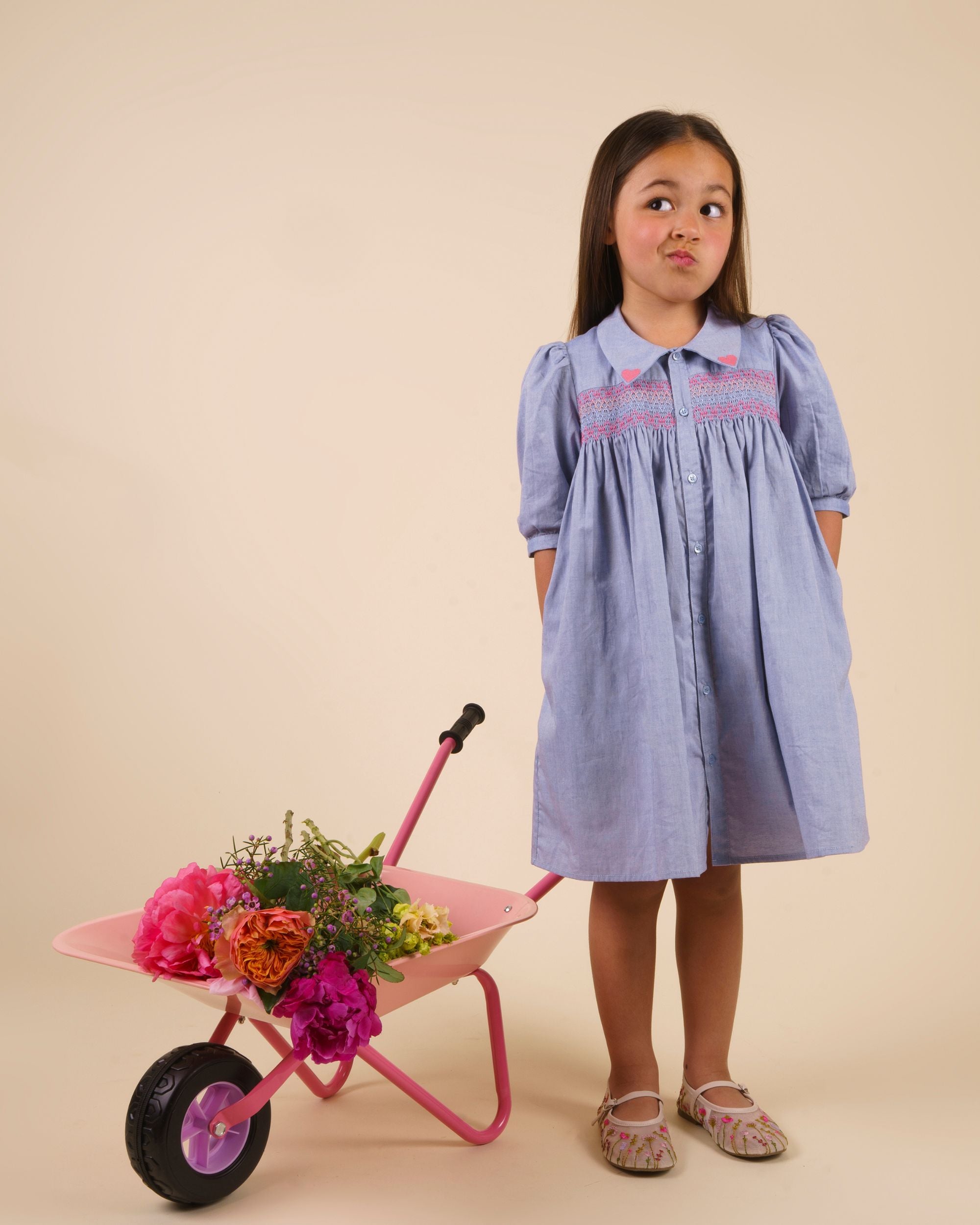 Early Sunday, Jane Dress in Summer Sky color, made from 100% cotton chambray fabric, featuring Smocked / embroidered yoke, peter pan collar and puffed short sleeves, shown in a model standing facing front wearing