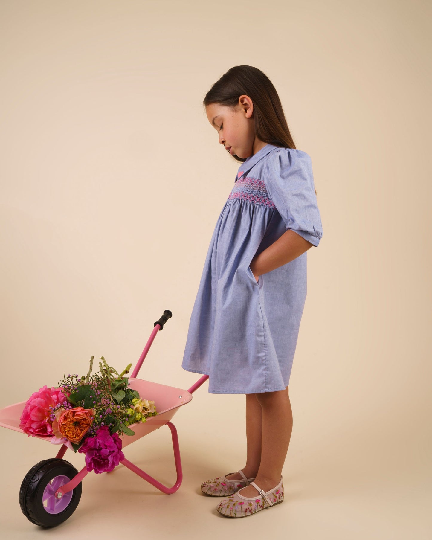 Early Sunday, Jane Dress in Summer Sky color, made from 100% cotton chambray fabric, featuring Smocked / embroidered yoke, peter pan collar and puffed short sleeves, shown in a side view of model wearing it