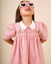 Early Sunday, Jane Dress in Peony color, made from 100% cotton gingham checks fabric, featuring Smocked / embroidered yoke, peter pan collar and puffed short sleeves, shown in a close up shot of model wearing it