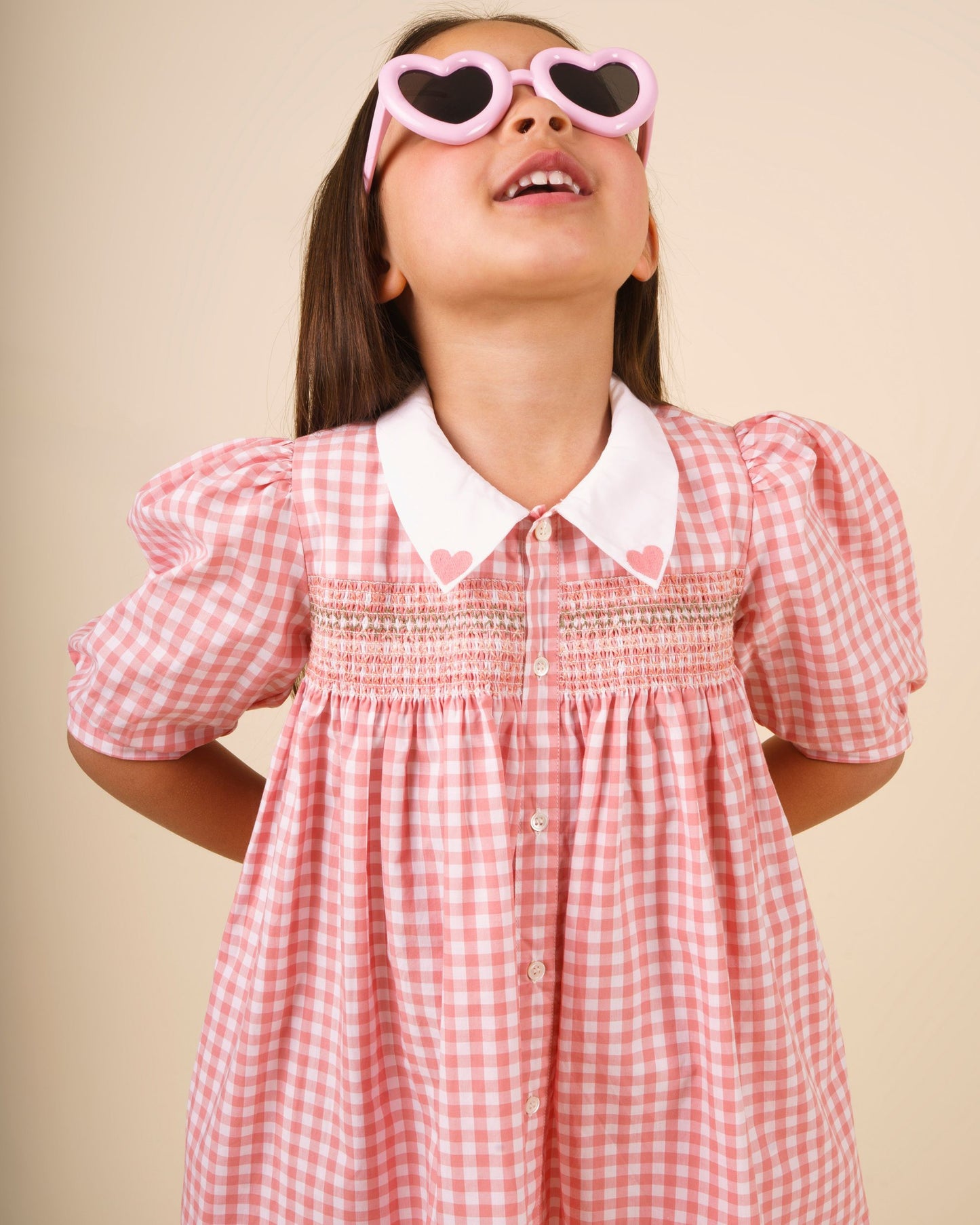 Early Sunday, Jane Dress in Peony color, made from 100% cotton gingham checks fabric, featuring Smocked / embroidered yoke, peter pan collar and puffed short sleeves, shown in a close up shot of model wearing it