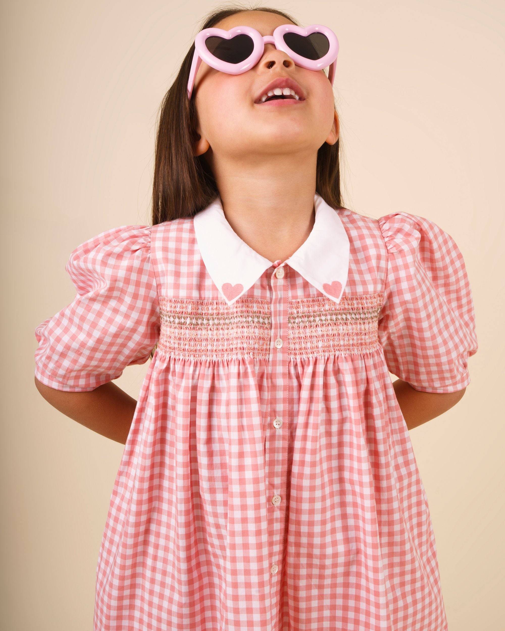 Early Sunday, Jane Dress in Peony color, made from 100% cotton gingham checks fabric, featuring Smocked / embroidered yoke, peter pan collar and puffed short sleeves, shown in a close up shot of model wearing it