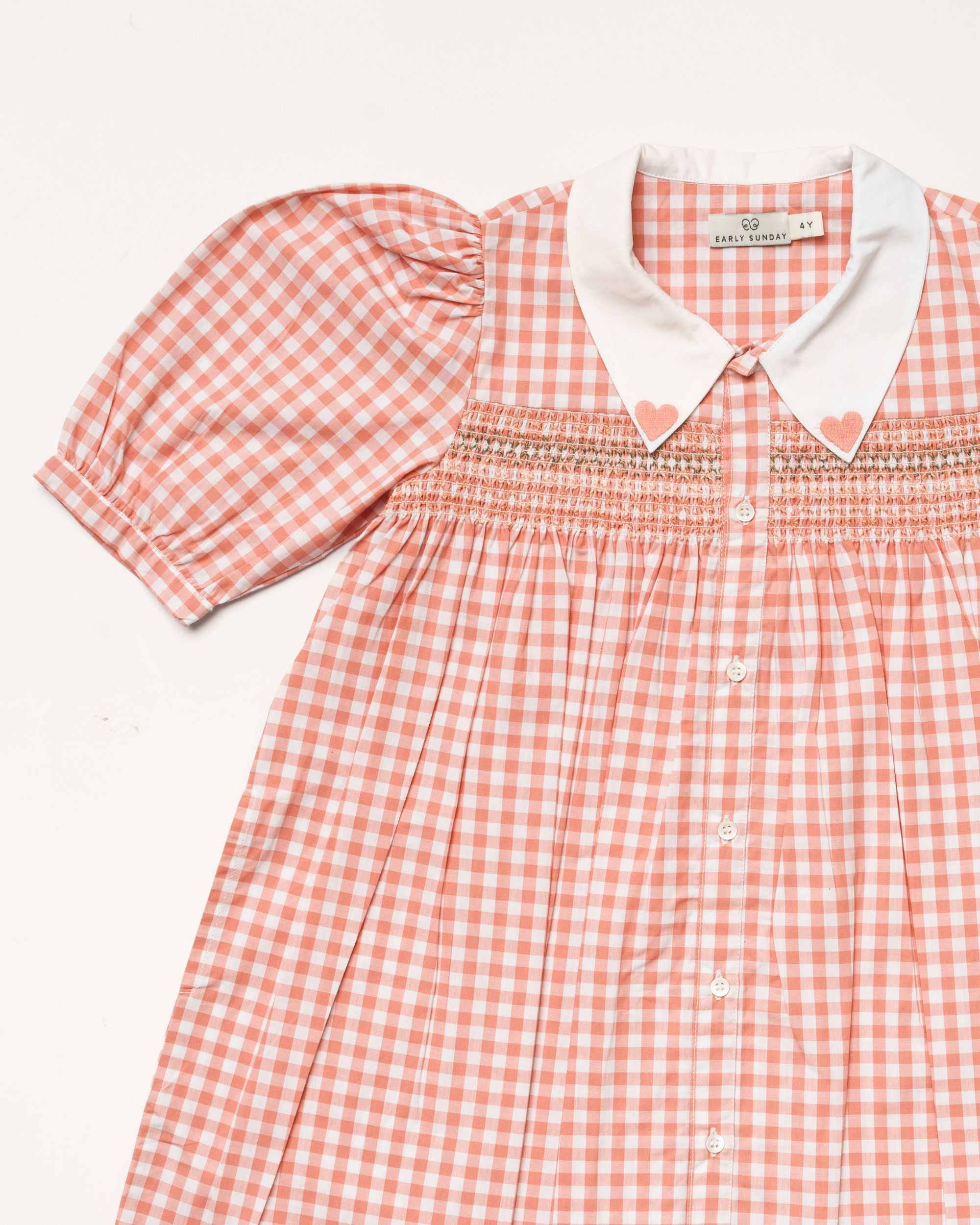 Early Sunday, Jane Dress in Peony color, made from 100% cotton gingham checks fabric, featuring Smocked / embroidered yoke, peter pan collar and puffed short sleeves, shown in a closeup flat-lay view