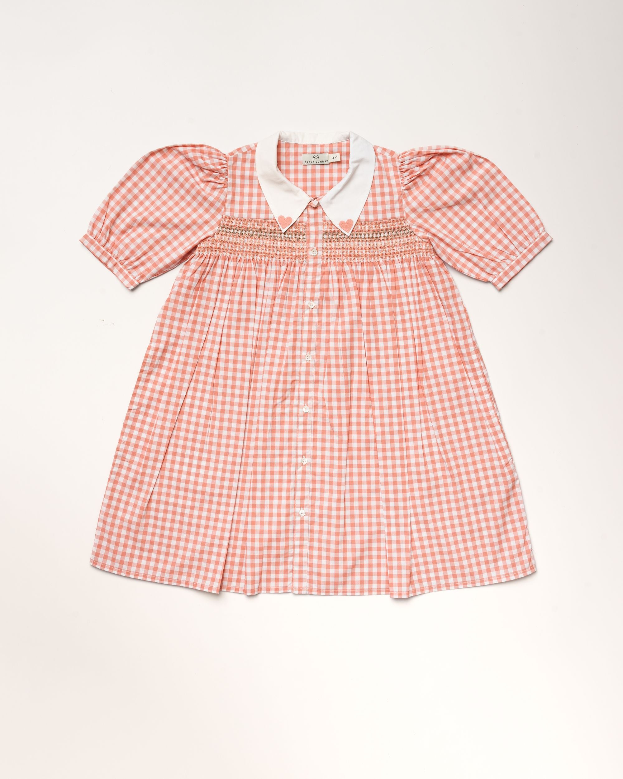 Early Sunday, Jane Dress in Peony color, made from 100% cotton gingham checks fabric, featuring Smocked / embroidered yoke, peter pan collar and puffed short sleeves, shown in a front flat-lay view