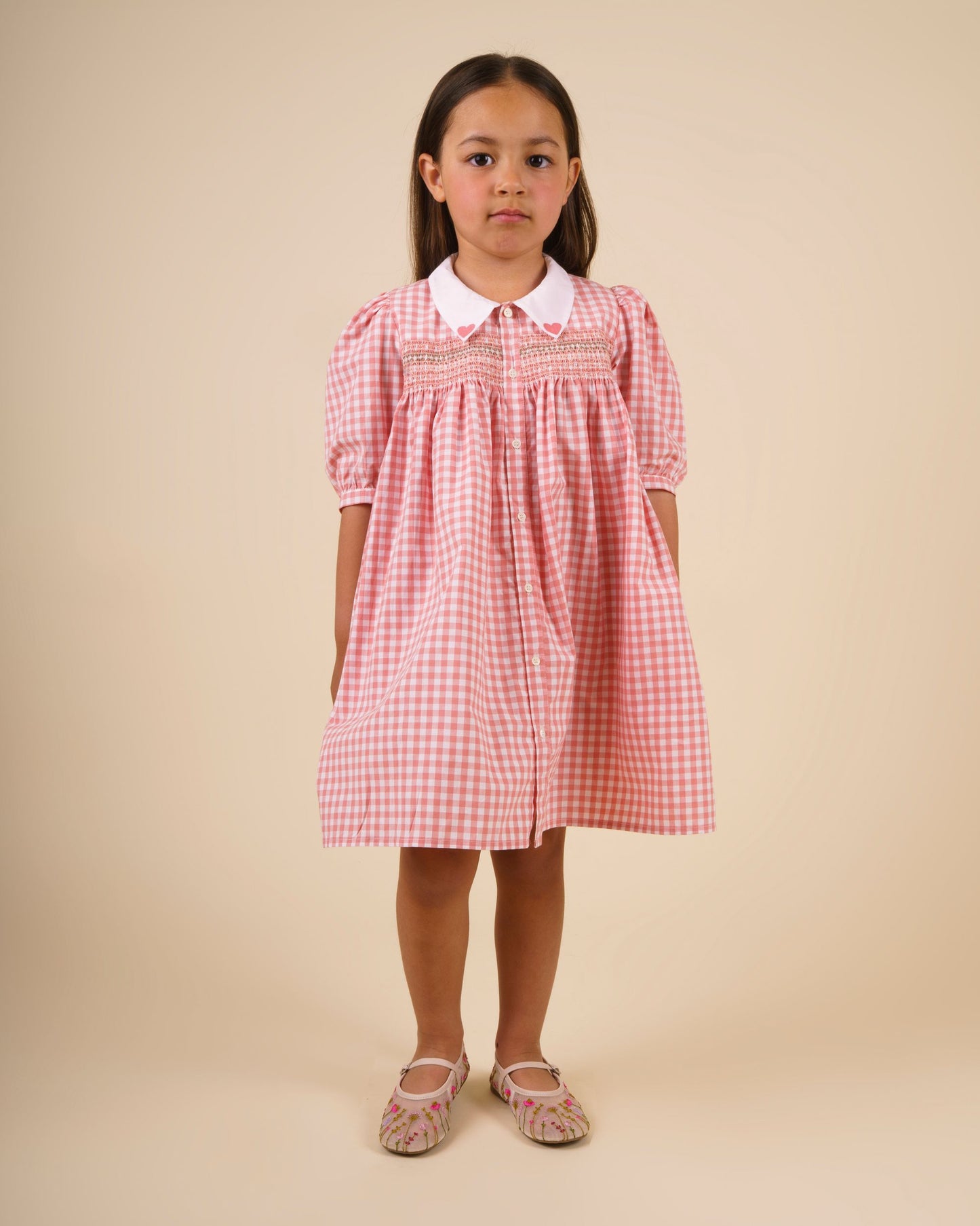 Early Sunday, Jane Dress in Peony color, made from 100% cotton gingham checks fabric, featuring Smocked / embroidered yoke, peter pan collar and puffed short sleeves, shown in a front view of model wearing
