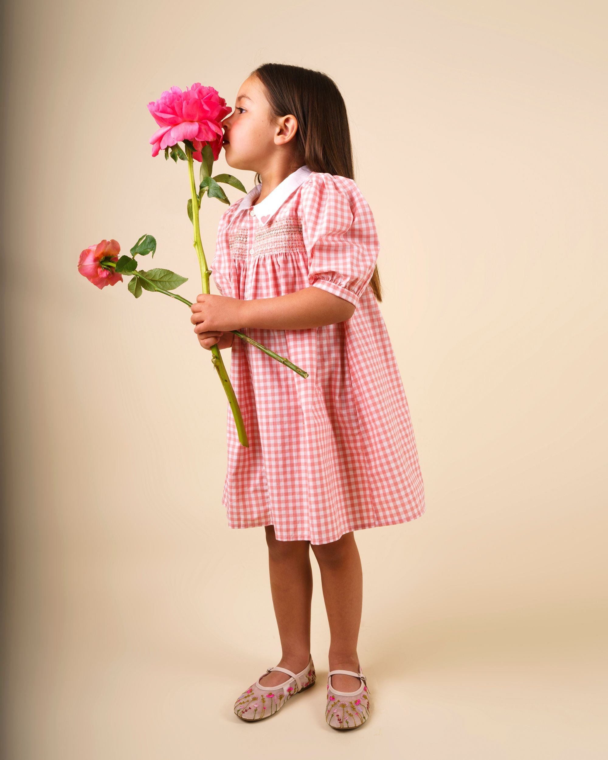 Early Sunday, Jane Dress in Peony color, made from 100% cotton gingham checks fabric, featuring Smocked / embroidered yoke, peter pan collar and puffed short sleeves, shown in a Model side pose / slight angle