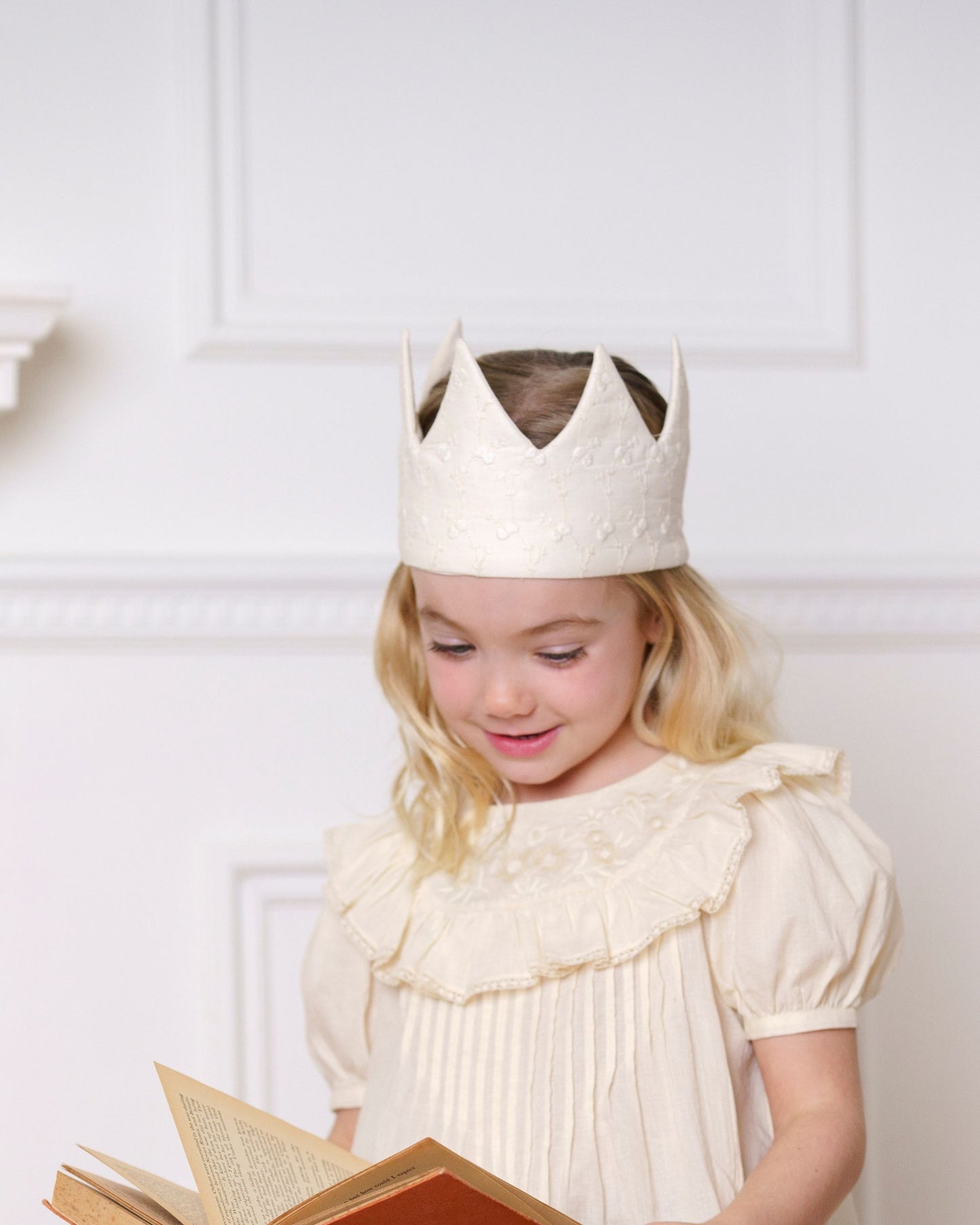 Early Sunday, Kids Crown in Hazelnut cream color, made from All over Embroidered Cotton Cambric fabric, featuring a design with pointed tips & an elasticated band, shown in a front view of a model wearing it in a studio setting