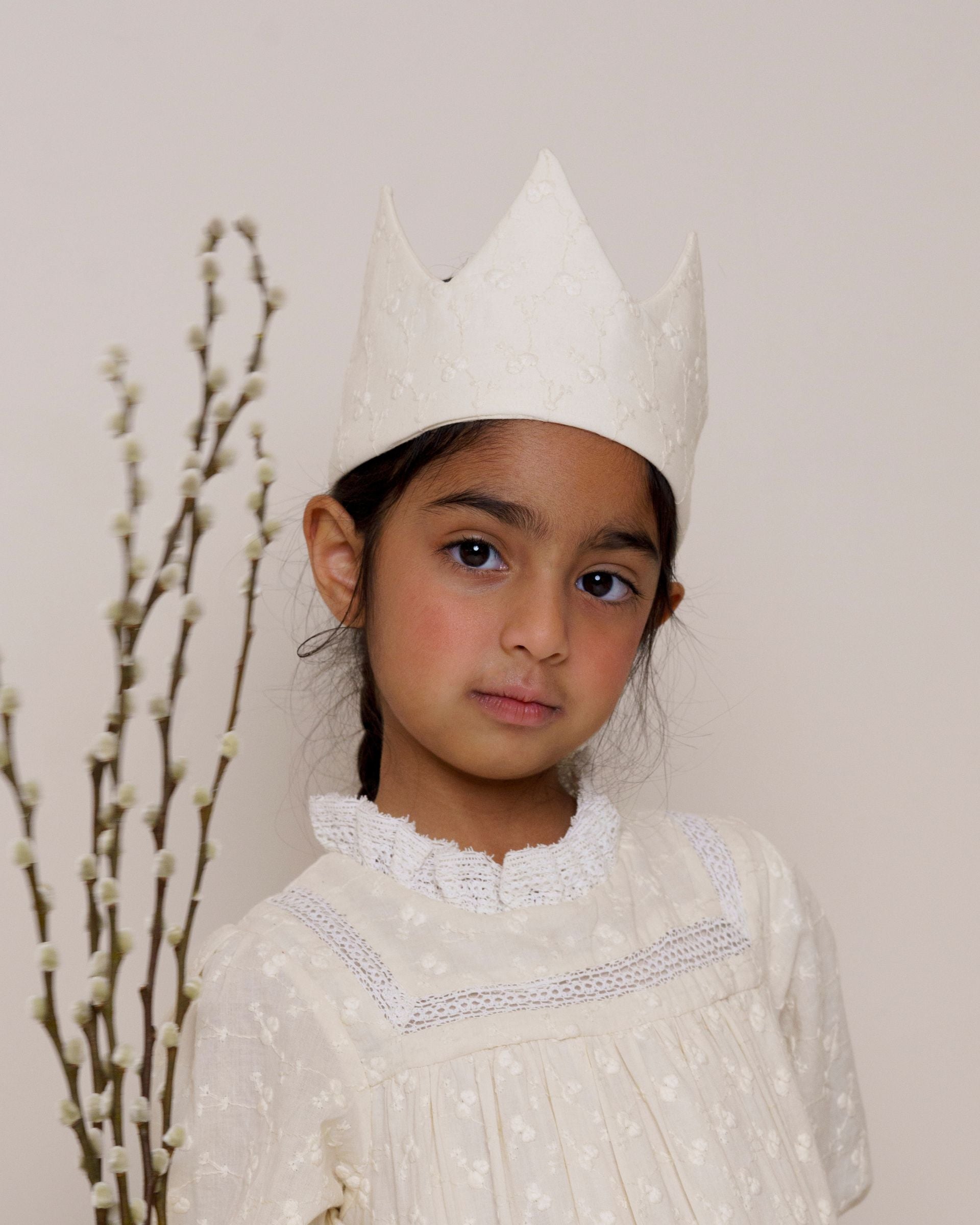 Early Sunday, Kids Crown in Hazelnut cream color, made from All over Embroidered Cotton Cambric fabric, featuring a design with pointed tips & an elasticated band, shown in an angled view of model wearing it