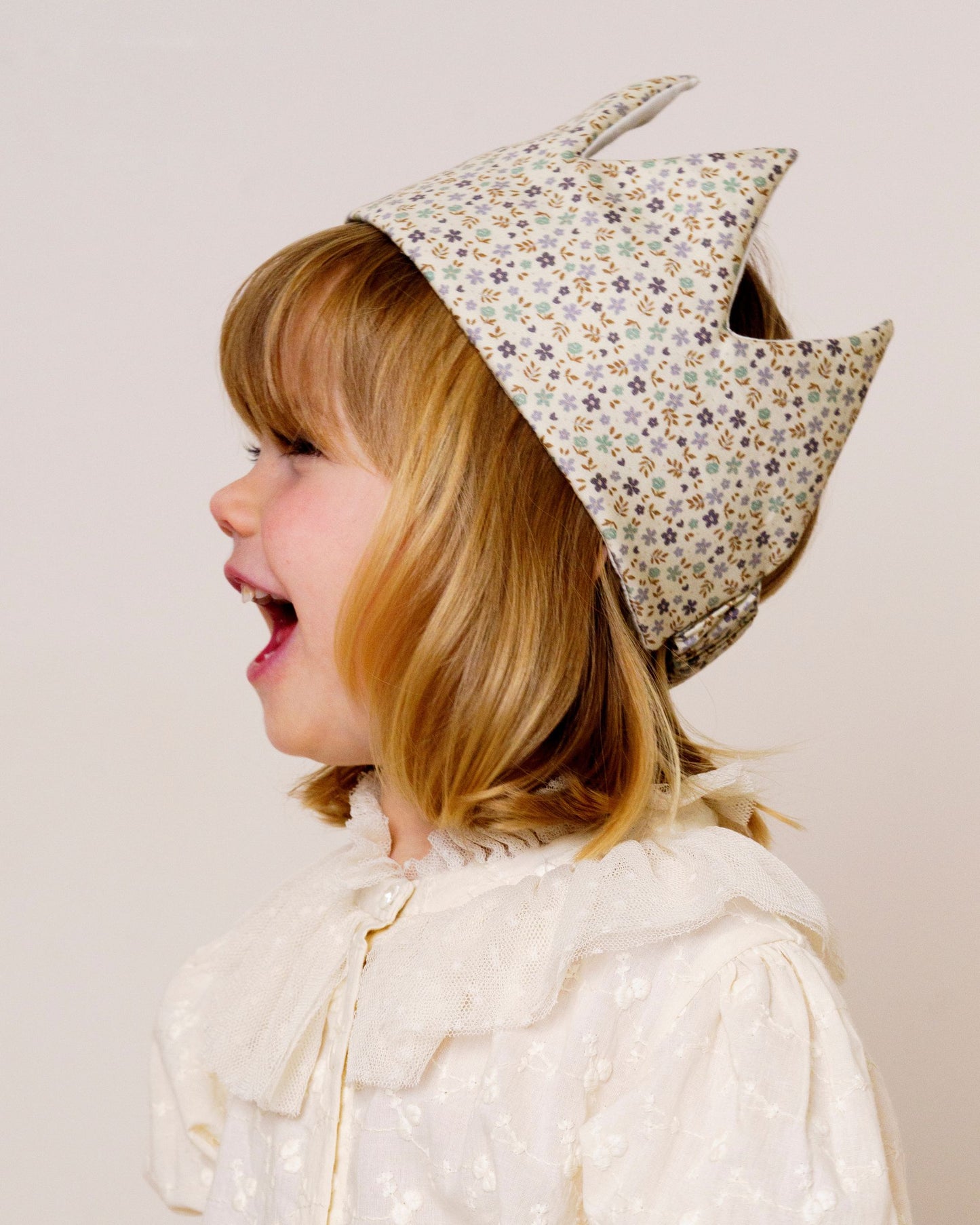 Early Sunday, Kids Crown in Mini Flowers print, made from Printed 60s Cotton Satin fabric, featuring a design with pointed tips & an elasticated band, shown in a close-up side view of a model wearing it