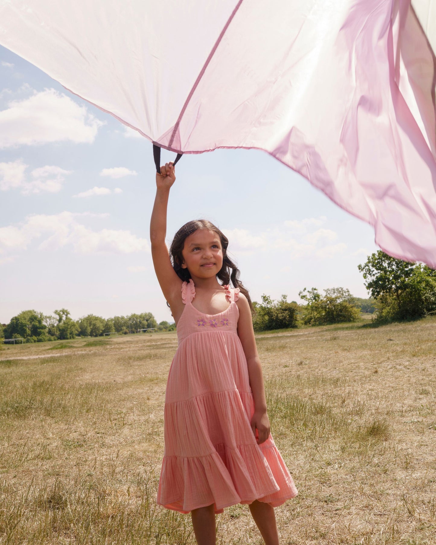 Early Sunday, Laura Dress in Candy Floss color, made from 100% cotton double gauze solid fabric, featuring Crisscross ruffle straps with delicate trim accents and a softly gathered tiered skirt, shown in a model wearing outdoors