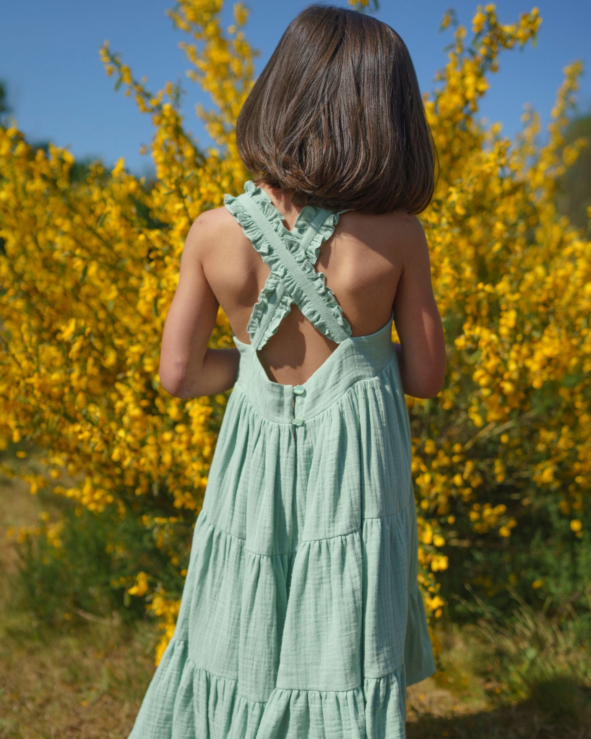 Early Sunday, Laura Dress in Mint color, made from 100% cotton double gauze solid fabric, featuring Crisscross ruffle straps with delicate trim accents and a softly gathered tiered skirt, shown in a back view of model wearing