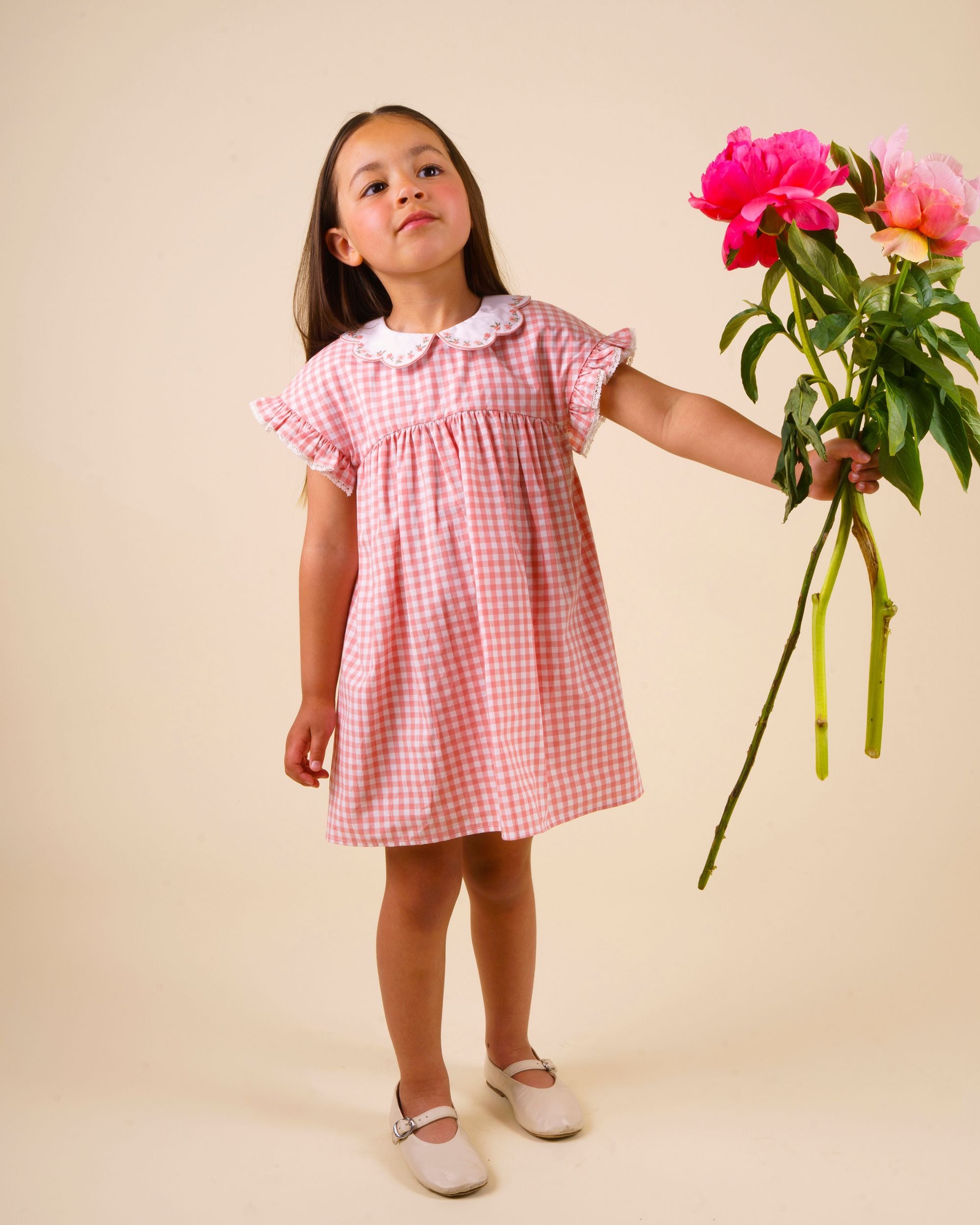Early Sunday, Lila Dress in Peony color, made from 100% cotton gingham checks fabric, featuring Scalloped neckline with contrast stitching, flutter sleeves, gentle gathers, and embroidered hem detail, shown in a model in motion