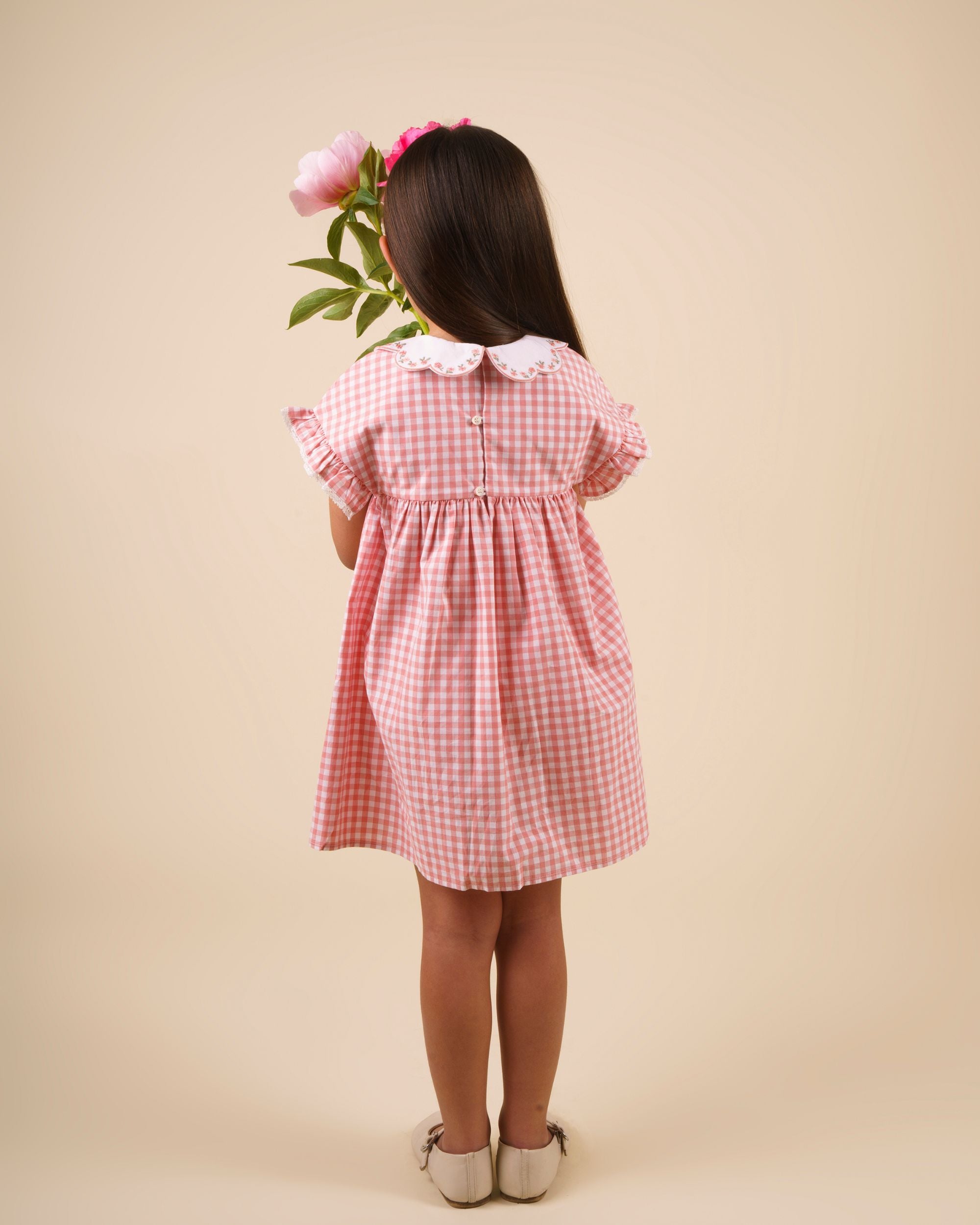 Early Sunday, Lila Dress in Peony color, made from 100% cotton gingham checks fabric, featuring Scalloped neckline with contrast stitching, flutter sleeves, gentle gathers, and embroidered hem detail, shown in a back view of model wearing
