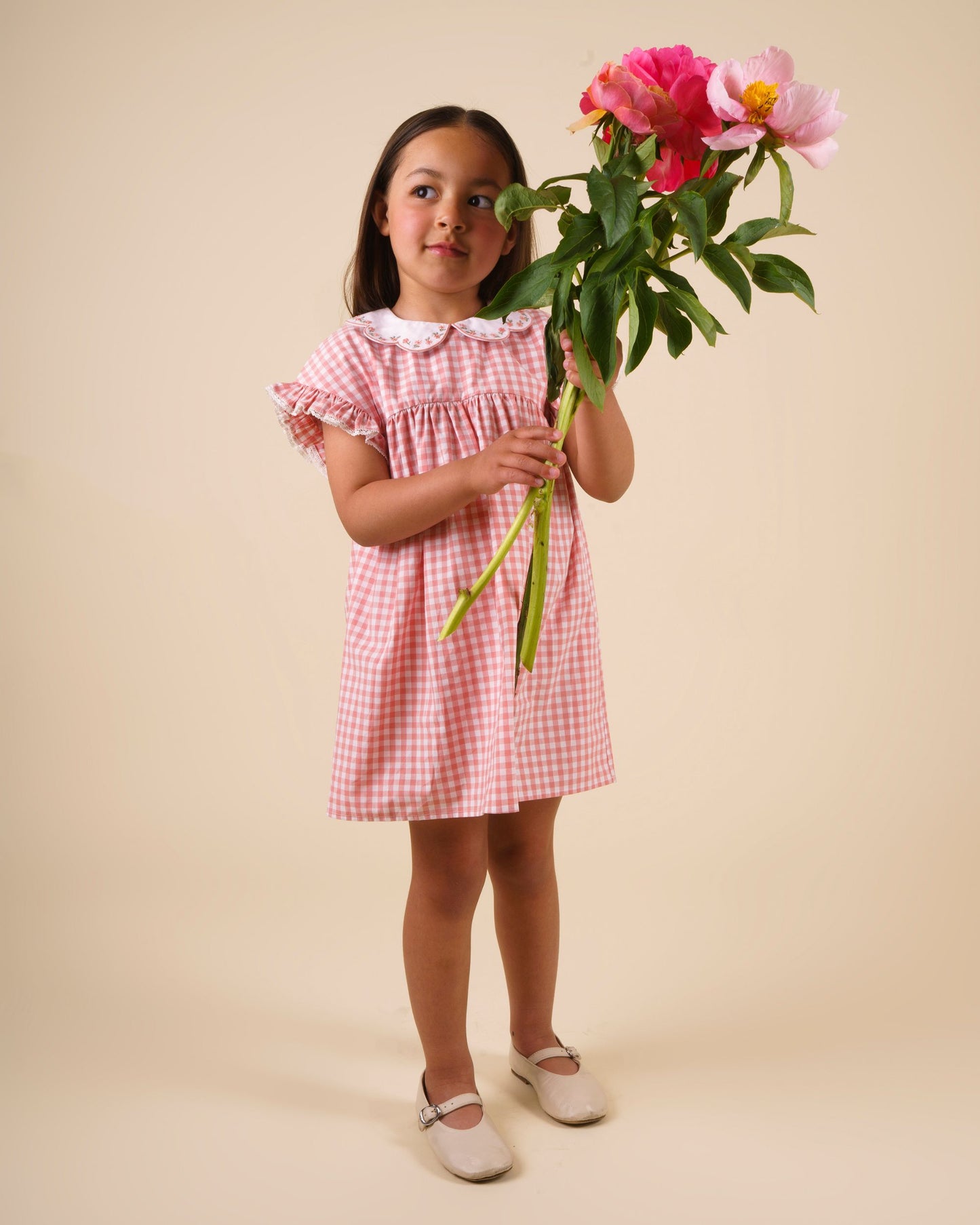 Early Sunday, Lila Dress in Peony color, made from 100% cotton gingham checks fabric, featuring Scalloped neckline with contrast stitching, flutter sleeves, gentle gathers, and embroidered hem detail, shown in a front view of model wearing