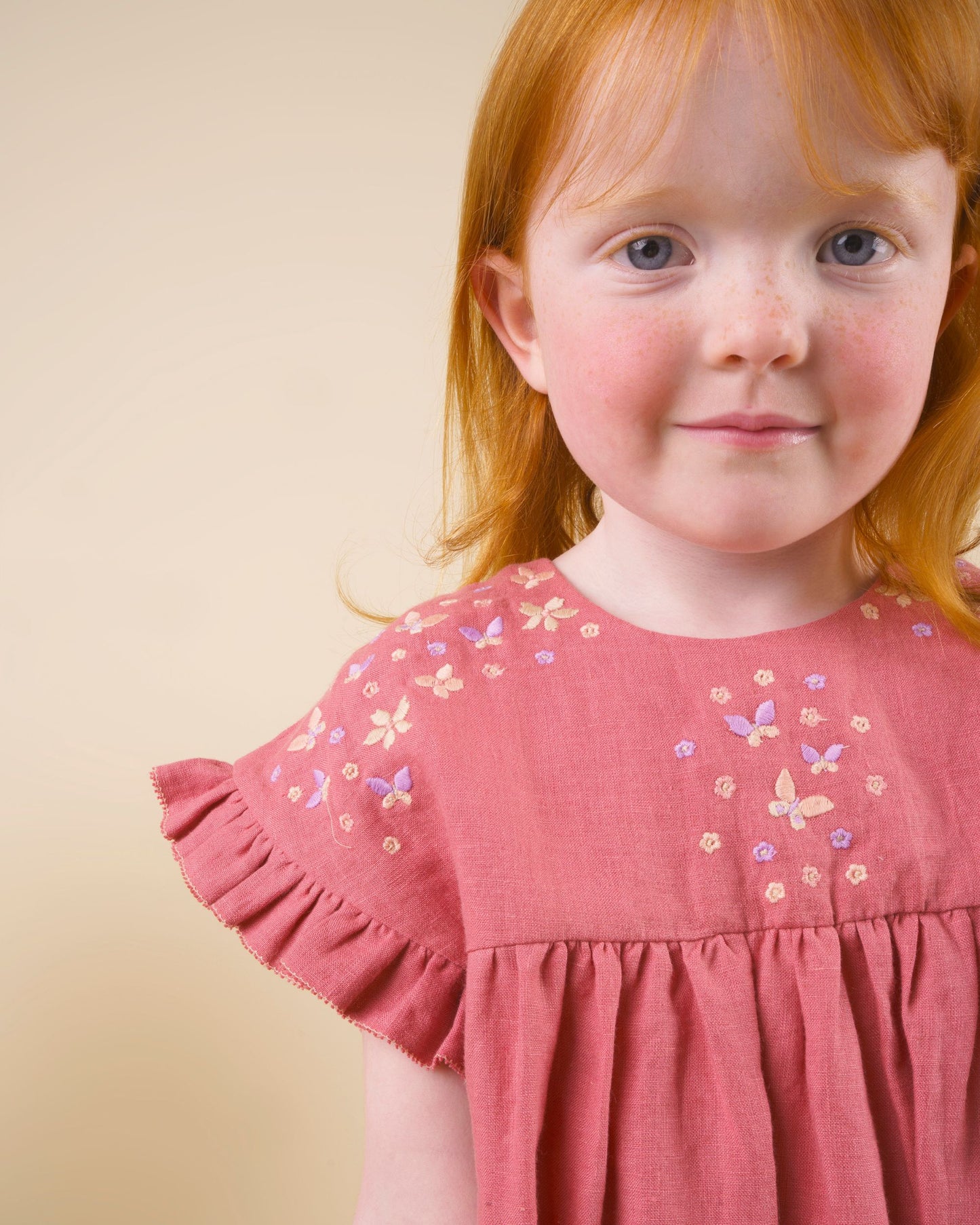 Early Sunday, Lila Dress in Peony color, made from 70% cotton 30% linen solid fabric, featuring Scalloped neckline with contrast stitching, flutter sleeves, gentle gathers, and embroidered hem detail, shown in a close up shot of model wearing it