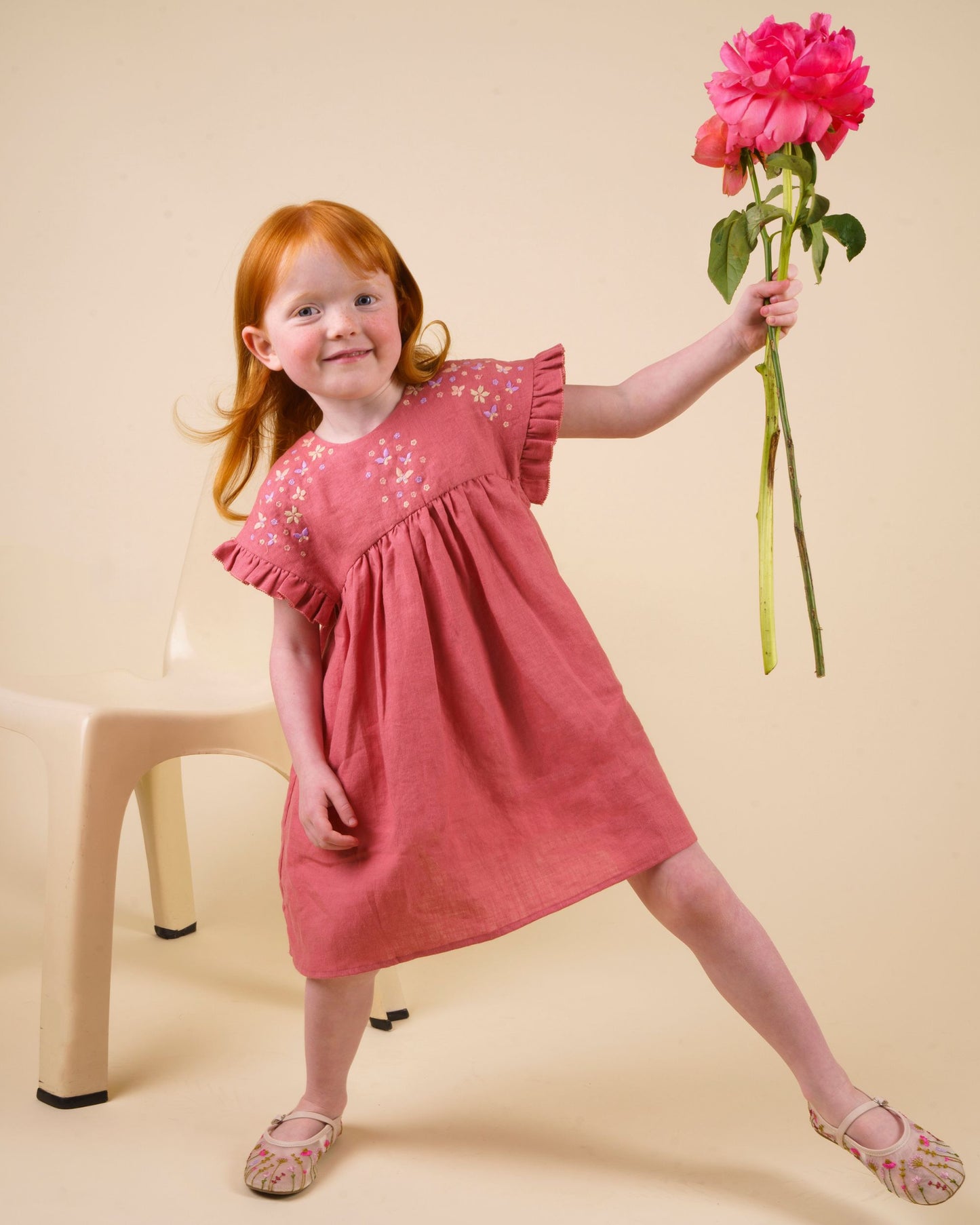 Early Sunday, Lila Dress in Peony color, made from 70% cotton 30% linen solid fabric, featuring Scalloped neckline with contrast stitching, flutter sleeves, gentle gathers, and embroidered hem detail, shown in a Model walking / movement shot