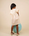 Early Sunday, Lila Dress in Sand color, made from 70% cotton 30% linen solid fabric, featuring Scalloped neckline with contrast stitching, flutter sleeves, gentle gathers, and embroidered hem detail, shown in a back view of model wearing