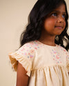 Early Sunday, Lila Dress in Sand color, made from 70% cotton 30% linen solid fabric, featuring Scalloped neckline with contrast stitching, flutter sleeves, gentle gathers, and embroidered hem detail, shown in a side view of model wearing