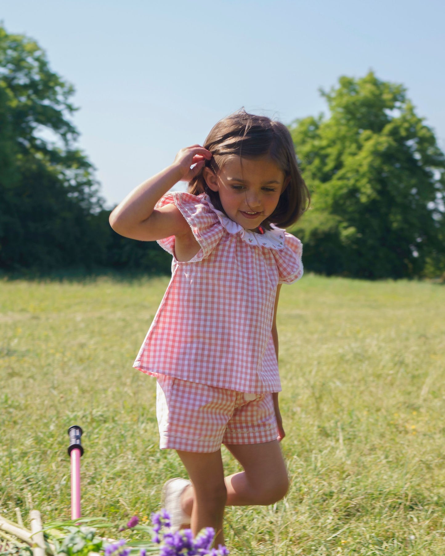 Early Sunday, Lola Blouse in Peony color, made from 100% cotton gingham checks fabric, featuring Ruffled neckline, flutter sleeves with contrast detailing, and gentle gathers for a sweet silhouette, shown in a model wearing outdoors