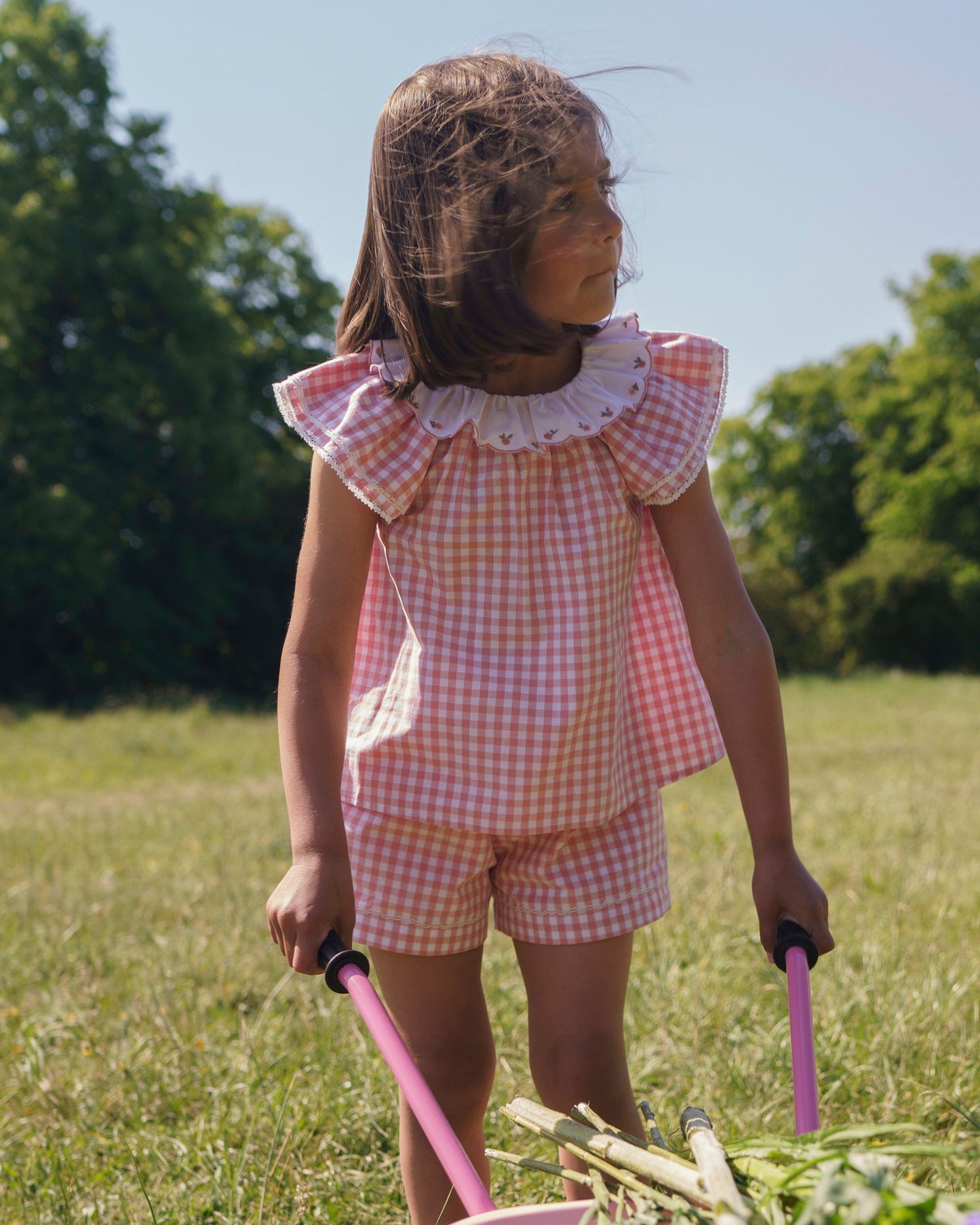 Early Sunday, Lola Blouse in Peony color, made from 100% cotton gingham checks fabric, featuring Ruffled neckline, flutter sleeves with contrast detailing, and gentle gathers for a sweet silhouette, shown in a model standing facing front wearing