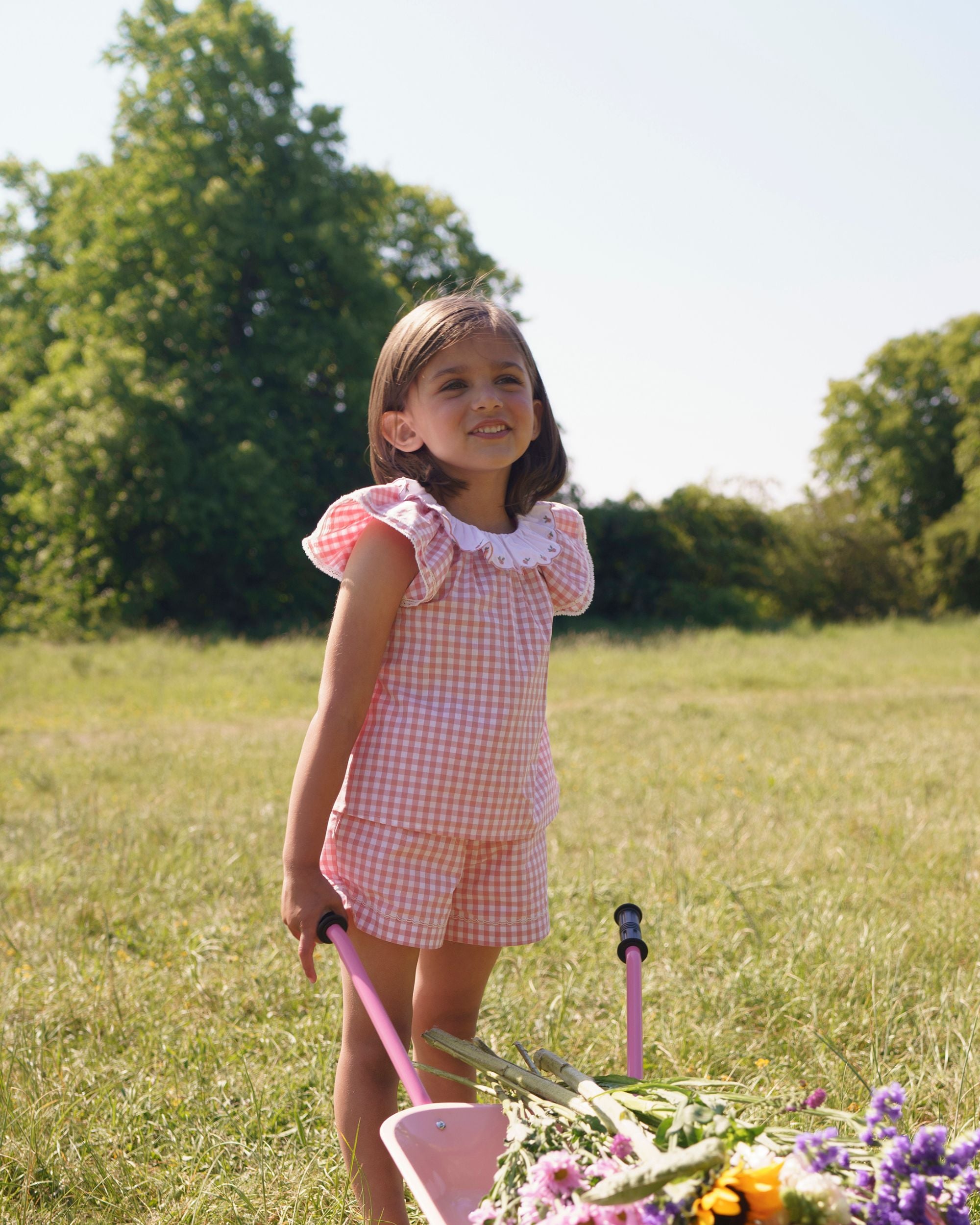 Early Sunday, Lola Blouse in Peony color, made from 100% cotton gingham checks fabric, featuring Ruffled neckline, flutter sleeves with contrast detailing, and gentle gathers for a sweet silhouette, shown in a Model walking / movement shot