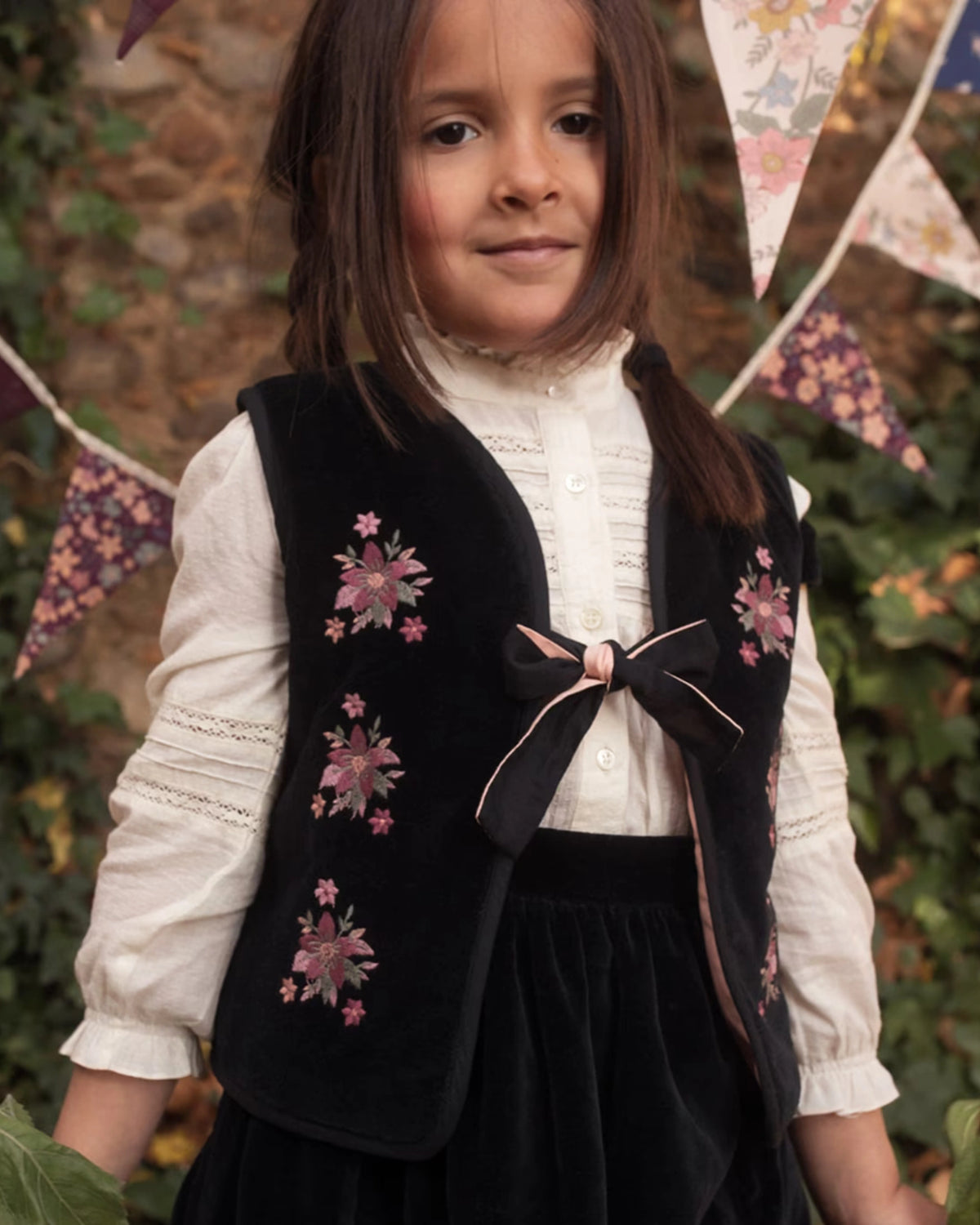 Early Sunday, Lou Vest jacket in Black, made from Cotton Velvet Velour's fabric, featuring sleeveless quilted silhouette with tie-up bow and floral embroidery, shown in a front view of model playing outdoors