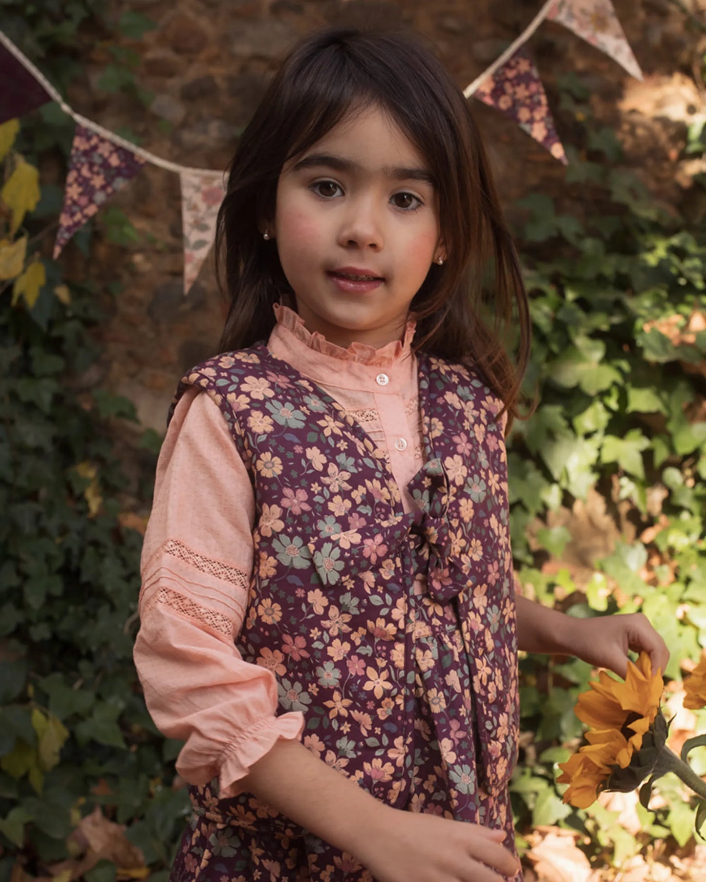 Early Sunday, Lou Vest jacket in Plum, made from Printed Cotton Satin fabric, featuring sleeveless quilted silhouette with tie-up bow detailing, shown in a front view of model playing outdoors