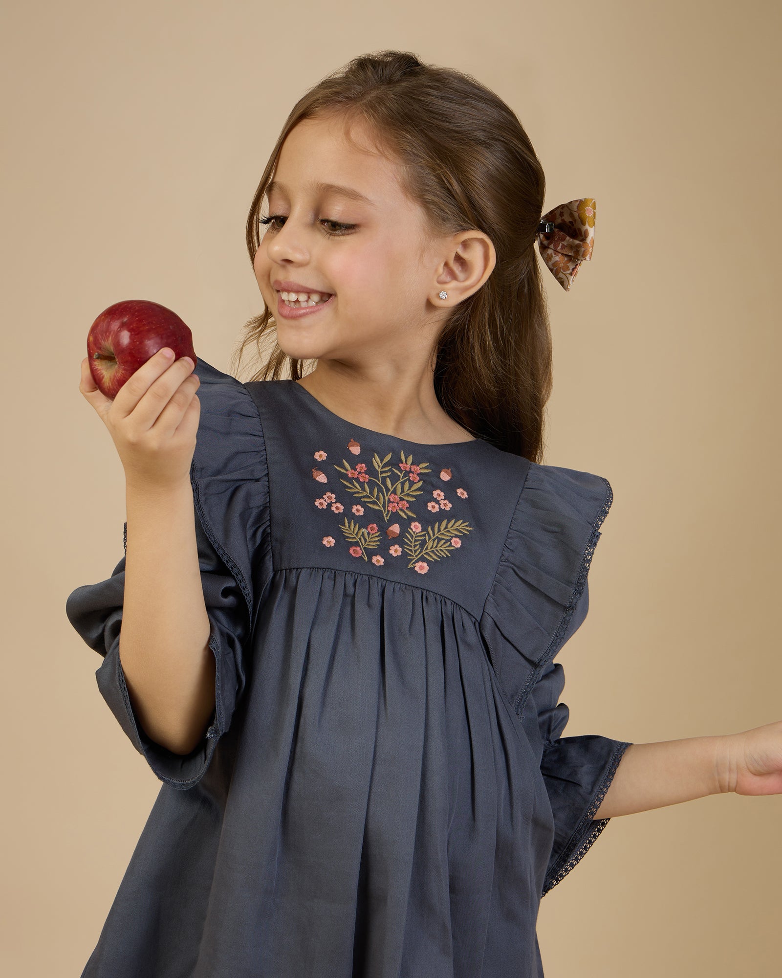 Early Sunday, Margot Dress in Stormy Blue, made from Cotton Satin fabric, featuring classic silhouette with floral embroidery around the yoke and ruffle detailing, shown in a front view of a model wearing it in a studio setting