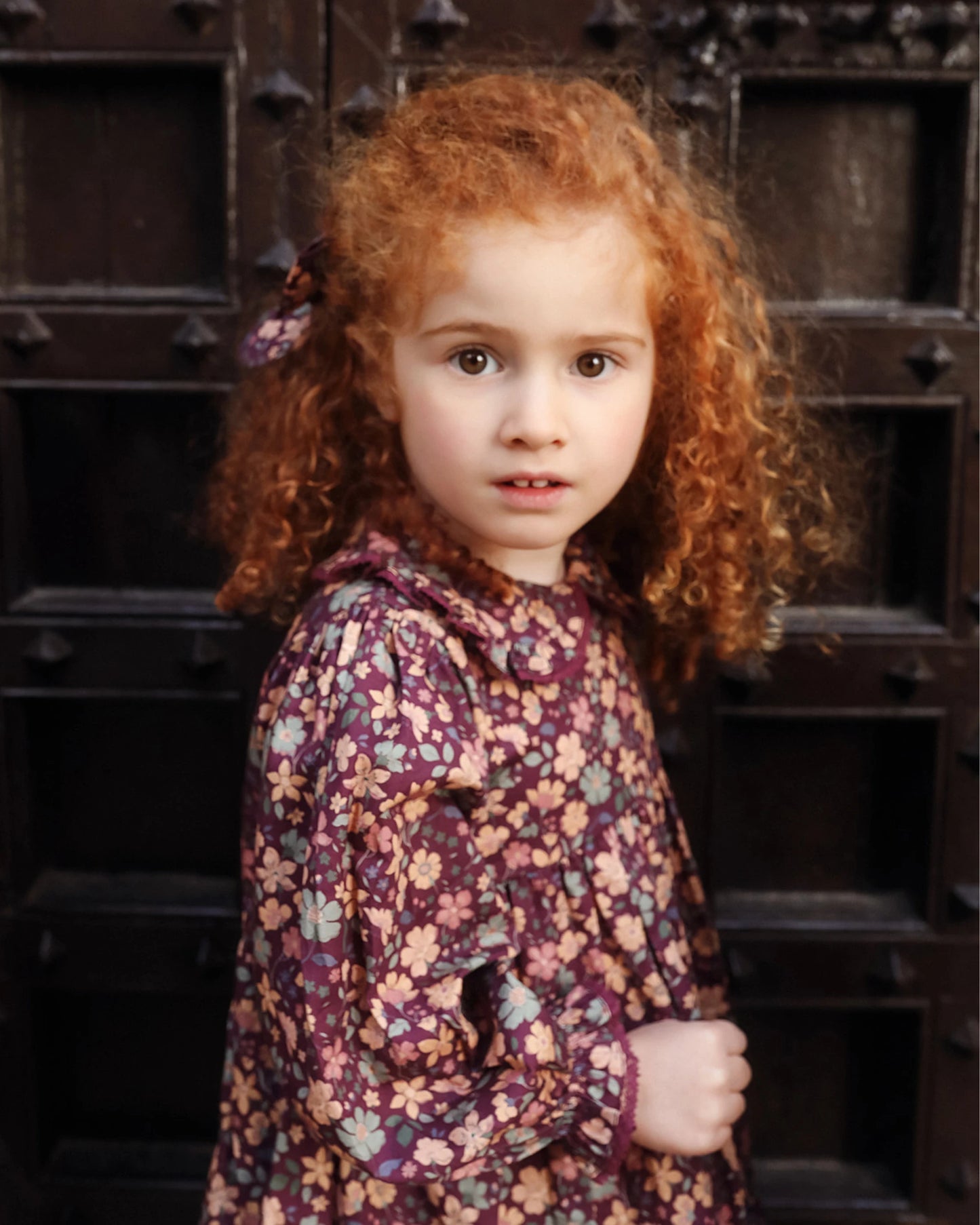 Early Sunday, Marguerite Dress in Garden Flowers Plum, made from Printed Cotton Satin fabric, featuring floral print and detailed lace yoke with Peter Pan collar, shown in a side view of model wearing it