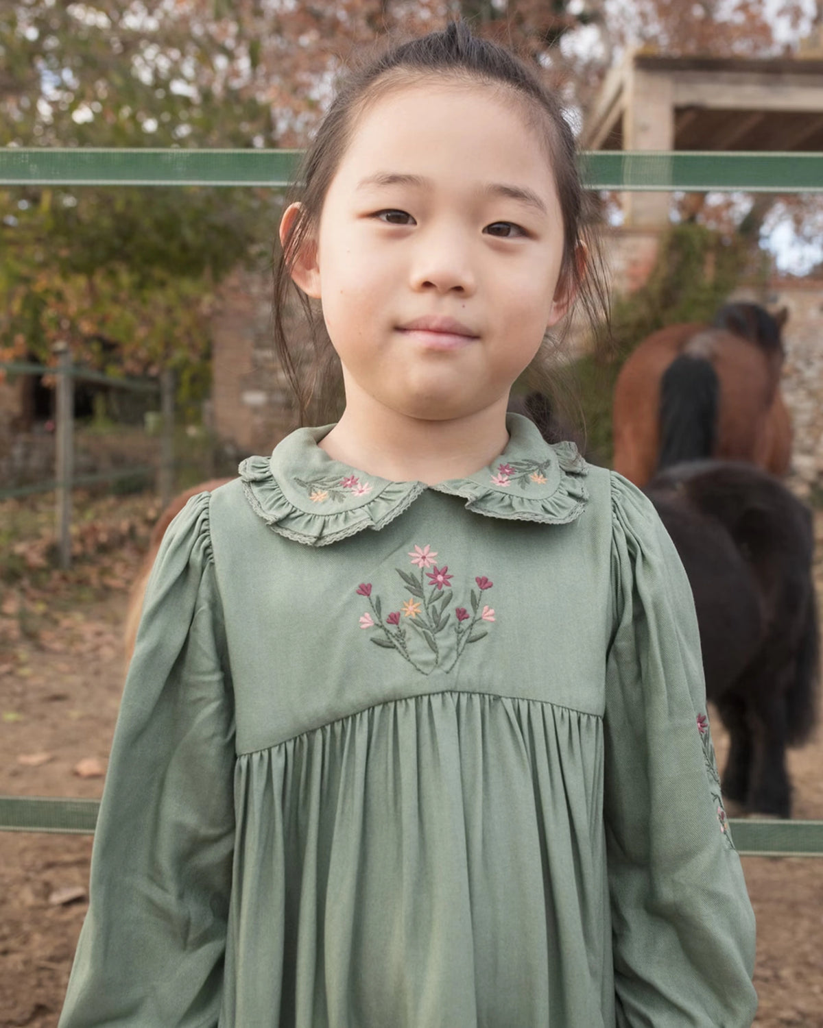 Early Sunday, Marguerite Dress in Sage Green, made from Viscose Drapey Twill fabric, featuring floral embroidery with frilled Peter Pan collar and gathered sleeves, shown in a close up shot of model wearing outdoors